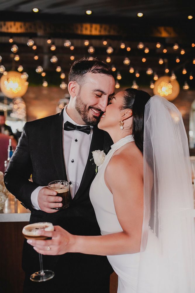 Newlyweds sharing intimate moment with champagne glasses under twinkling reception lights.