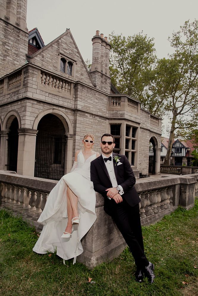 Wedding couple posing playfully in front of a gothic stone castle building with arched architecture.