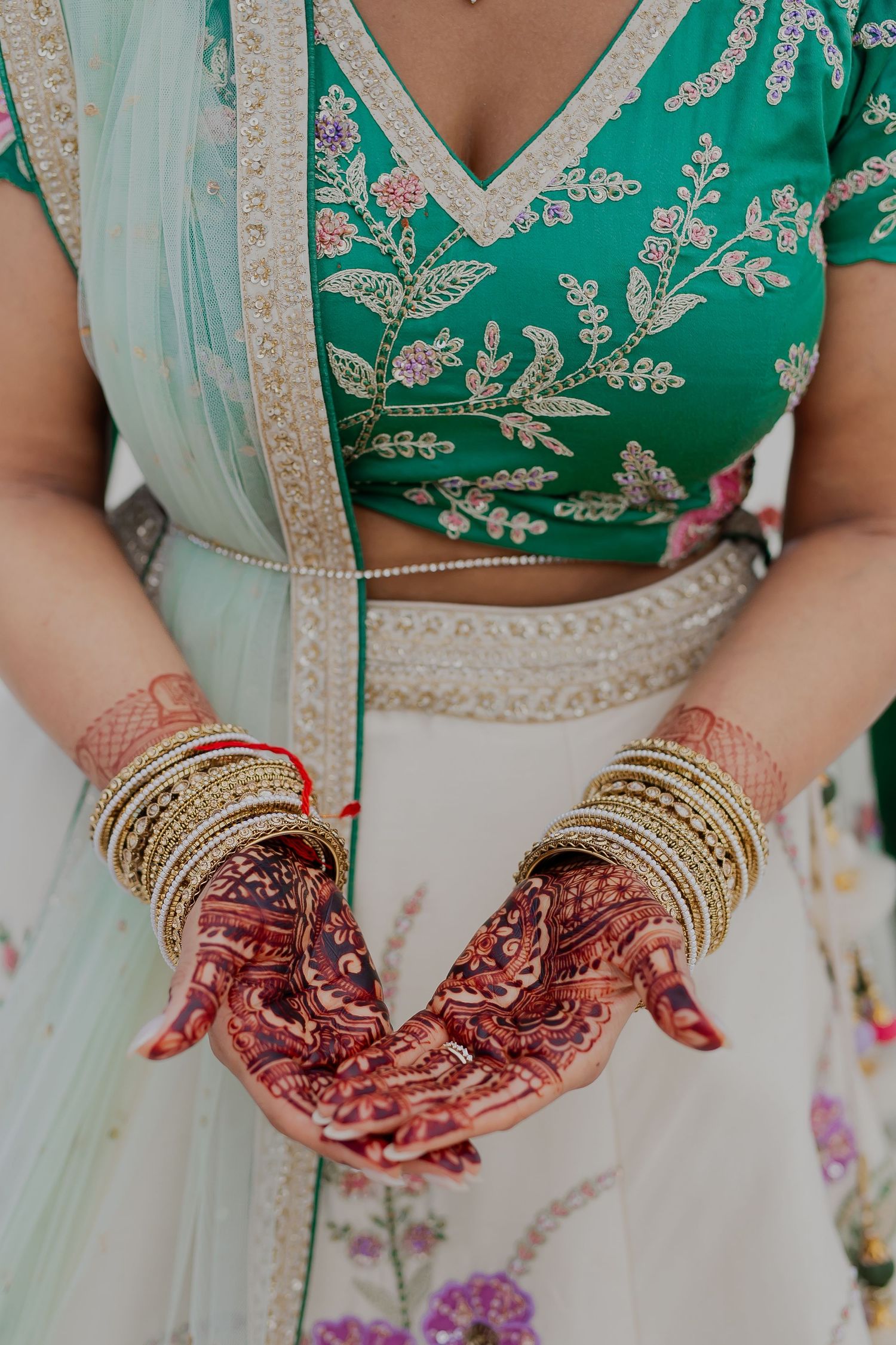 Close-up of henna adorned hands with gold bangles and embroidered green and white outfit.