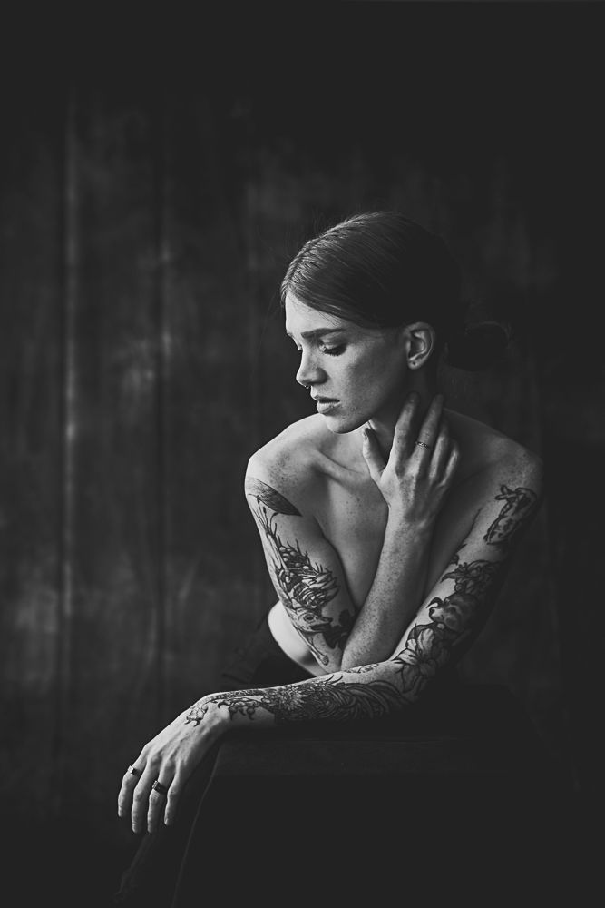 Artistic black and white portrait showing tattooed shoulders in contemplative pose against dark background.