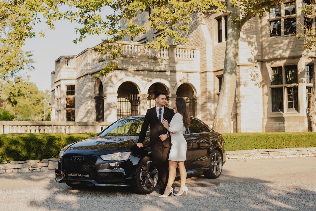A couple stands beside a black Audi car in front of an elegant stone mansion with arched architecture.
