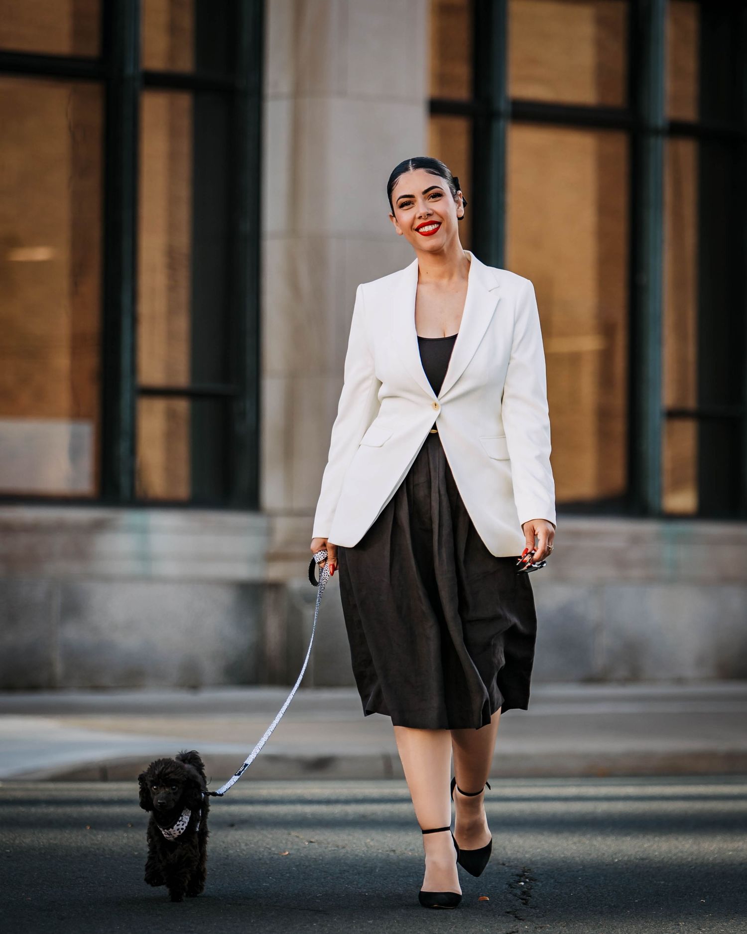 Woman in white blazer and black dress walks her small black dog on a leash outside an office building.