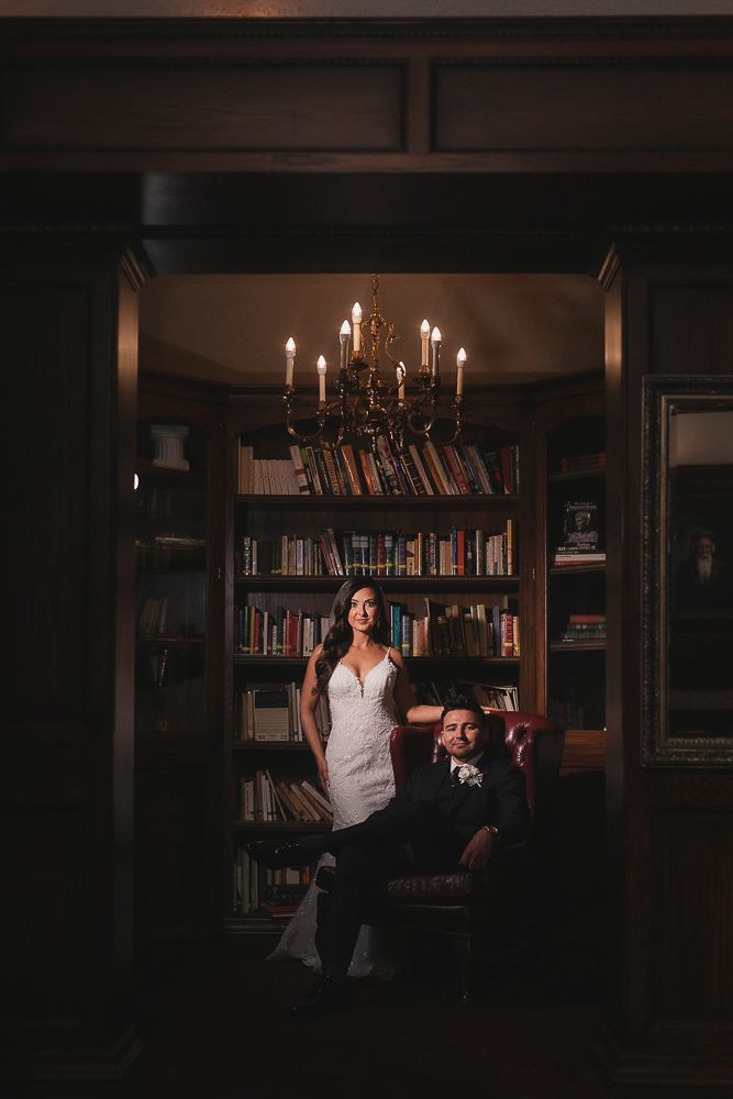 A couple poses in an elegant library setting with a chandelier and bookshelves creating a moody, romantic atmosphere.