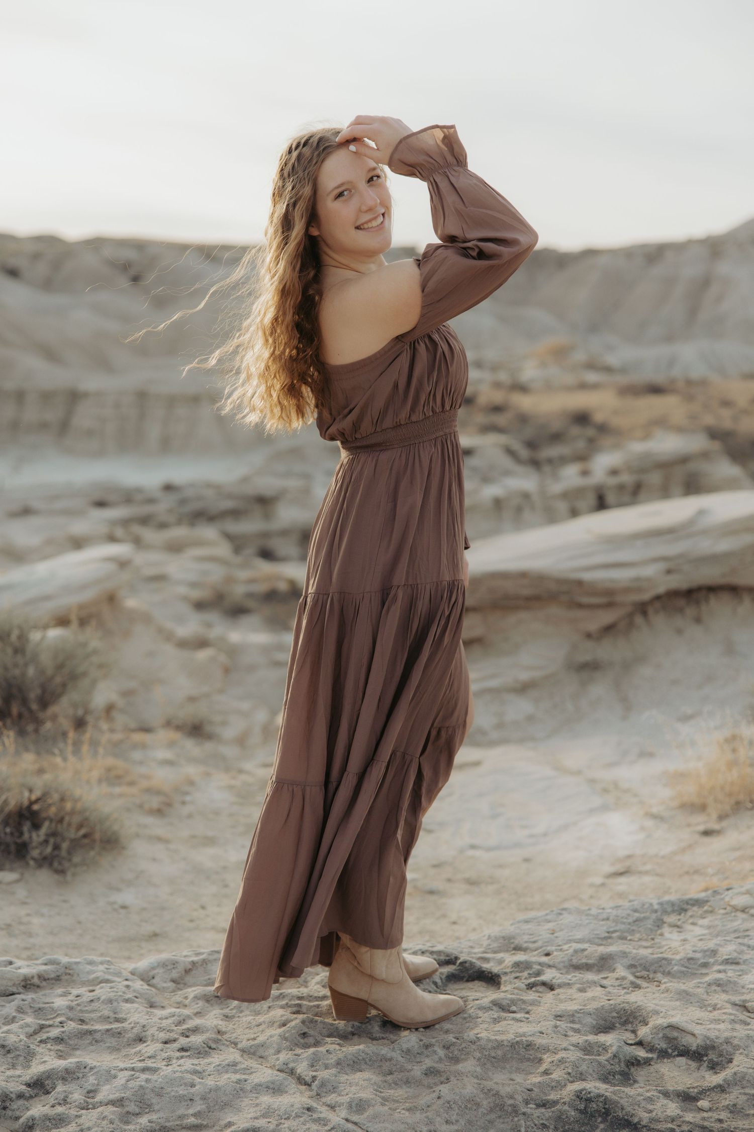 Woman in brown dress twirls playfully on rocky desert terrain.