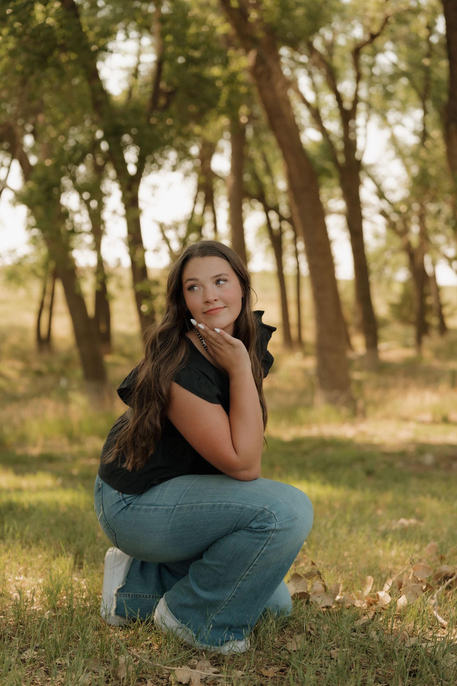 Young woman in black top and jeans crouching outdoors in a sunlit forest, posing thoughtfully with hand near chin.