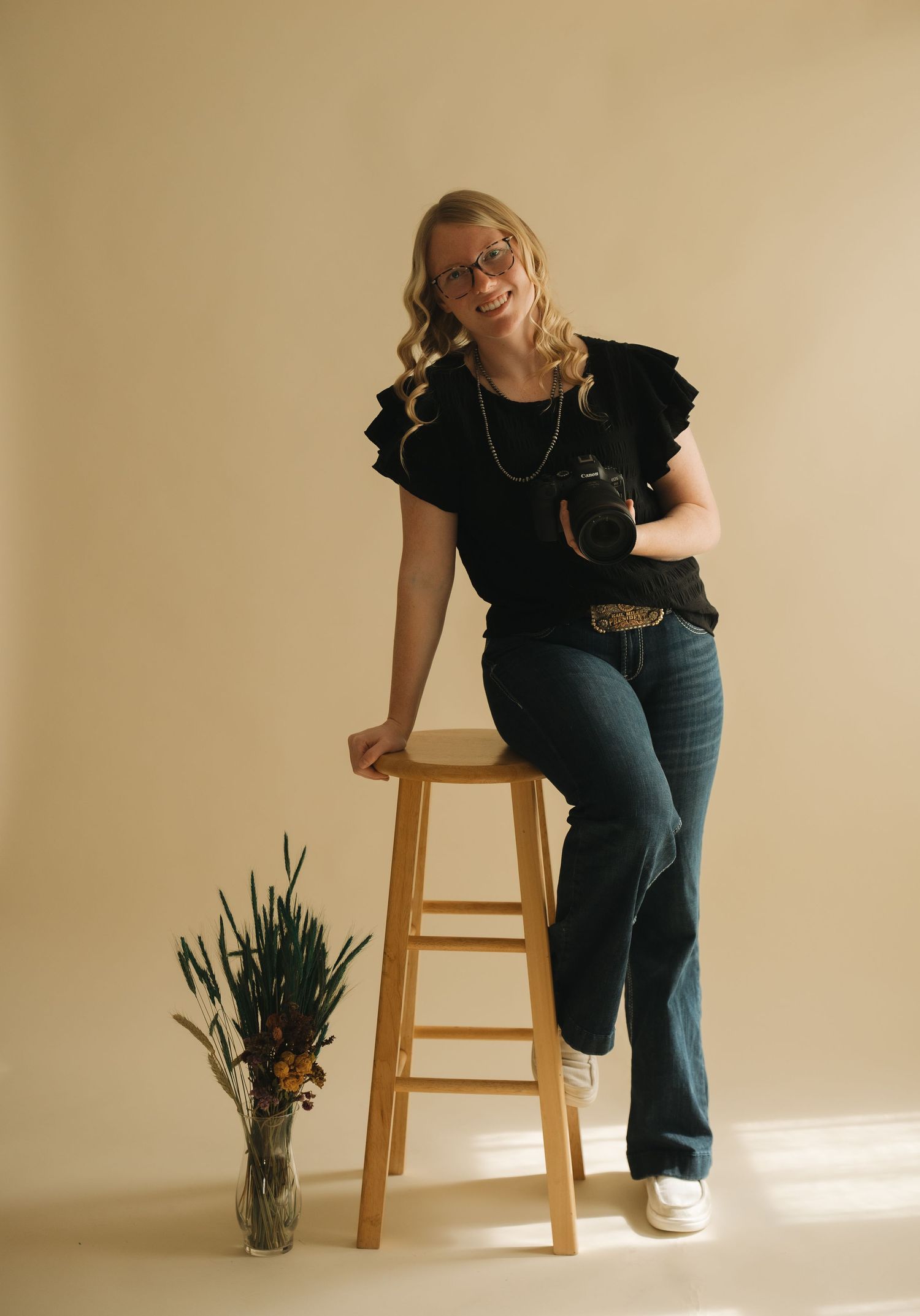 Portrait against beige wall with wooden stool and vase of flowers, wearing black top, jeans and white sneakers.