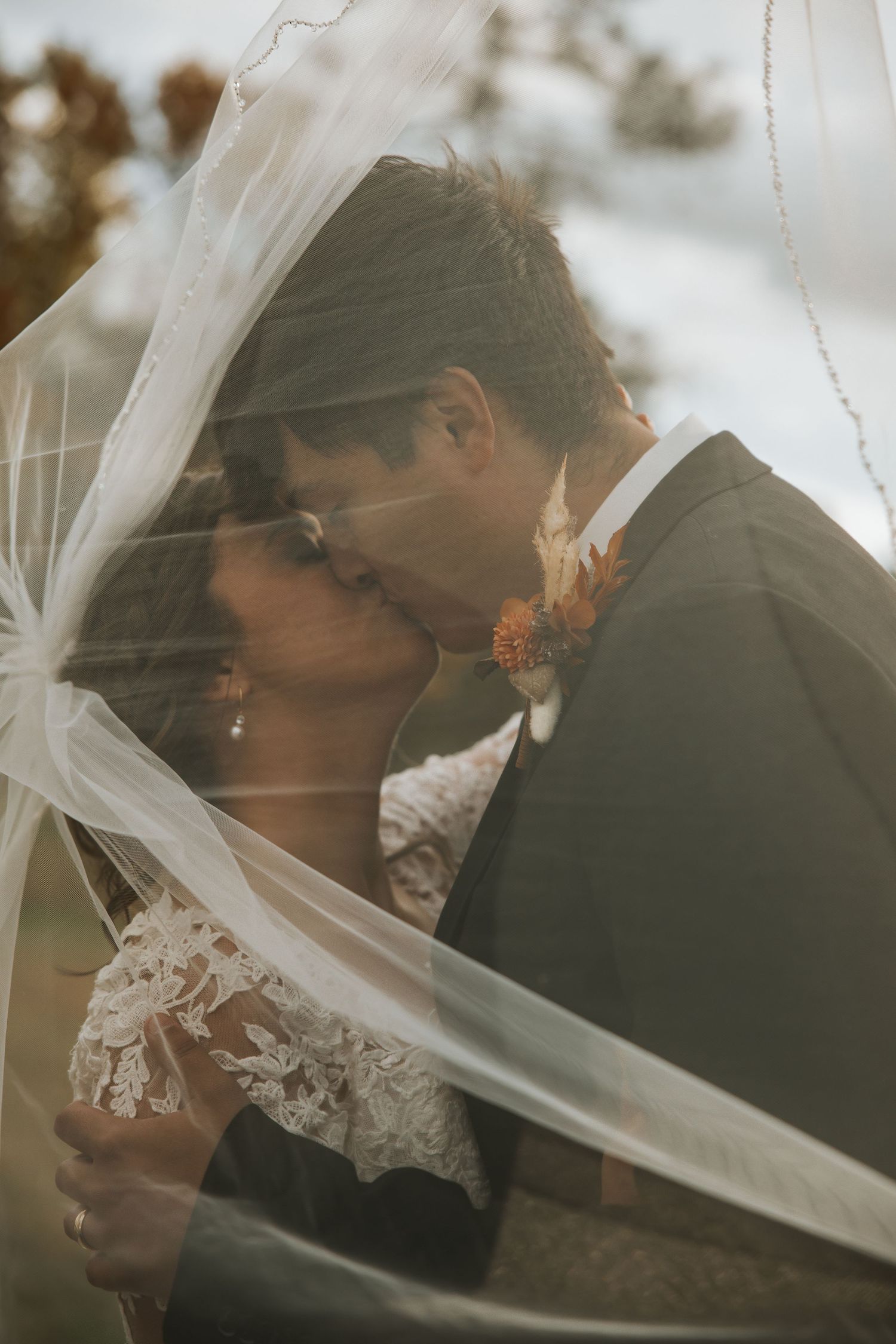 A romantic wedding photo showing a couple kissing under a delicate veil during their outdoor ceremony.