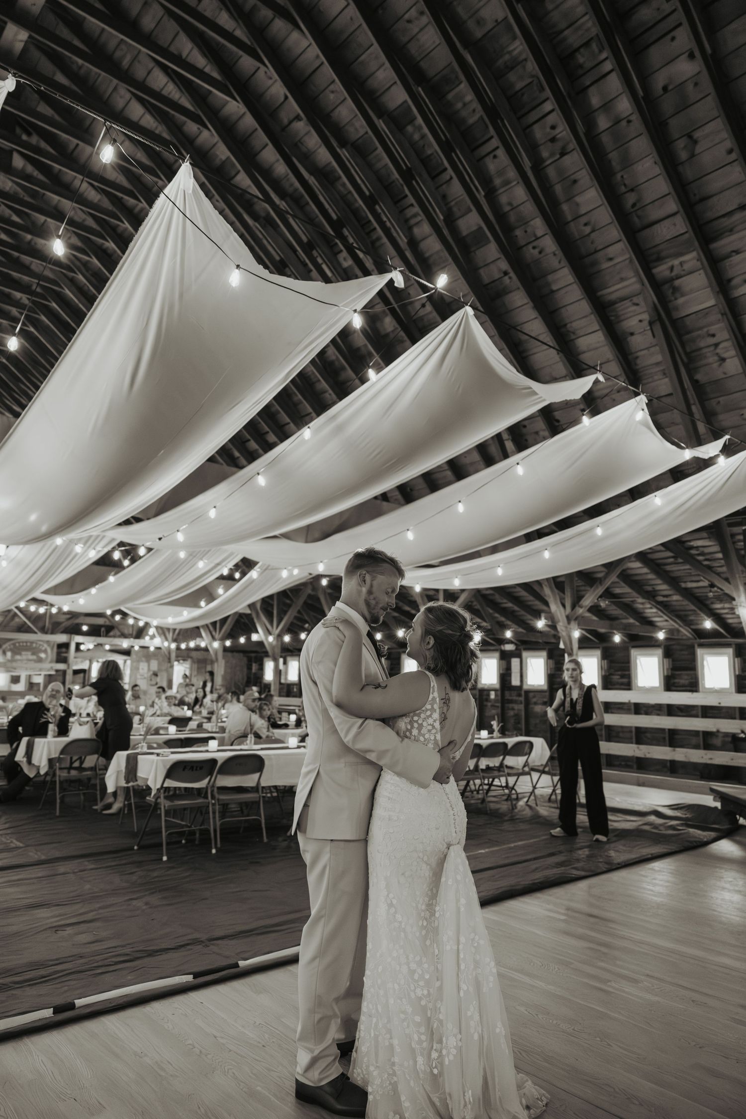 Black and white photo of a bride and groom sharing their first dance in a rustic barn venue with draped fabric ceiling.