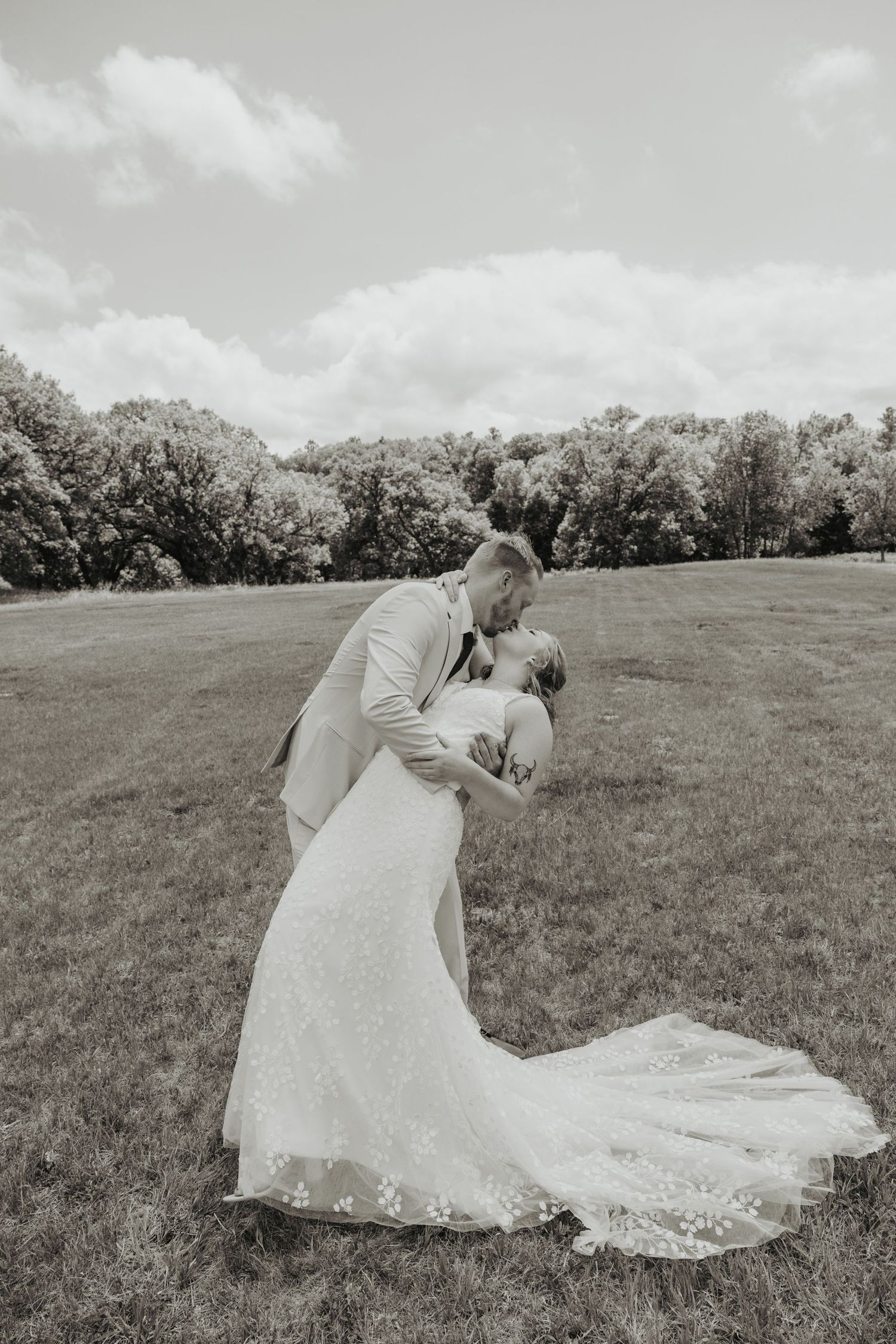Black and white photo of a groom dipping and kissing his bride in a lush green field on their wedding day.