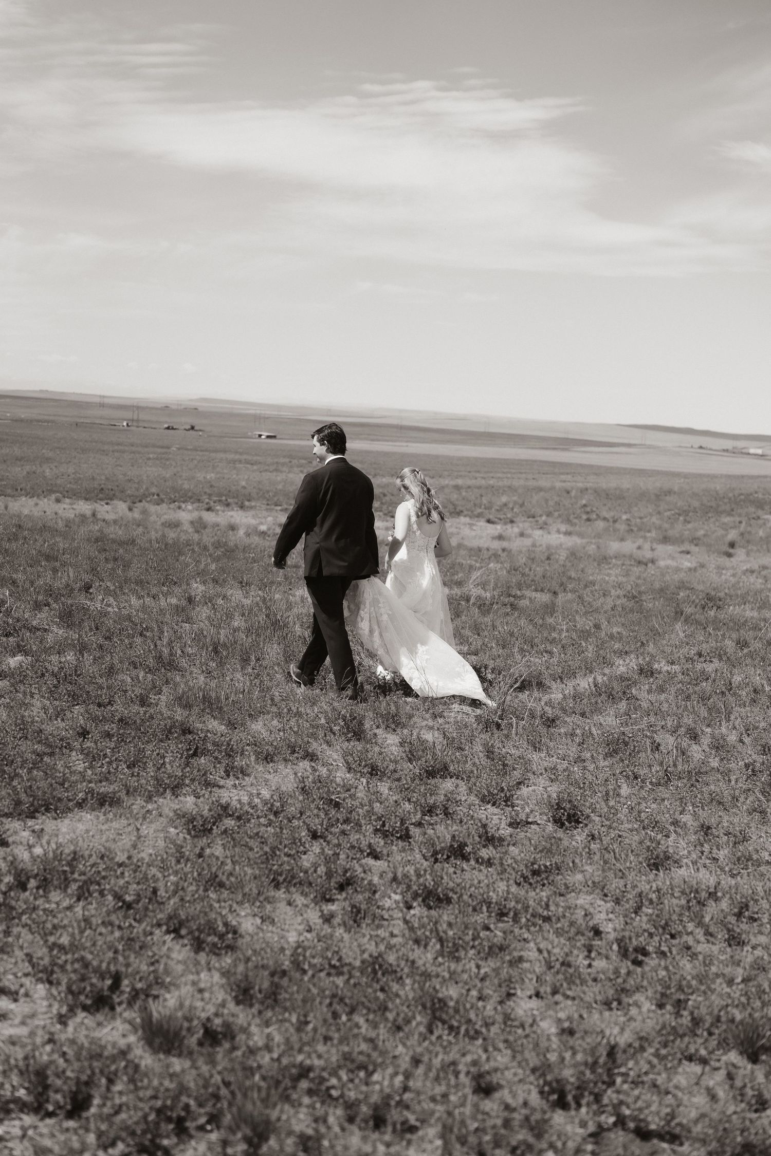 Black and white photo of a bride and groom walking through an open field, wedding dress flowing in the wind.