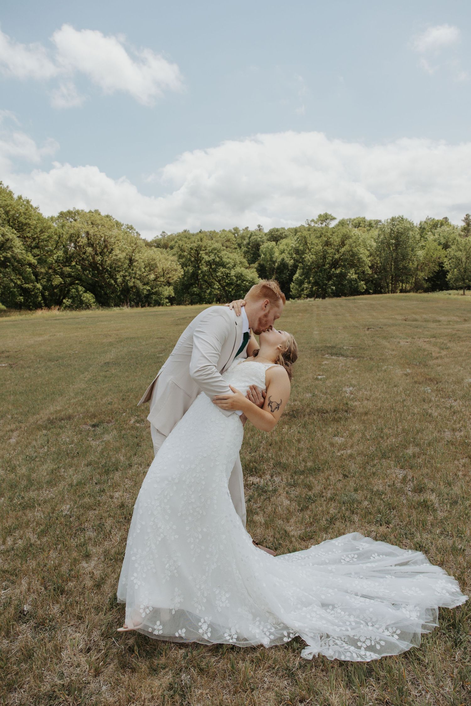 A groom in a tan suit dips his bride in a white lace gown for a romantic kiss in a lush green field.