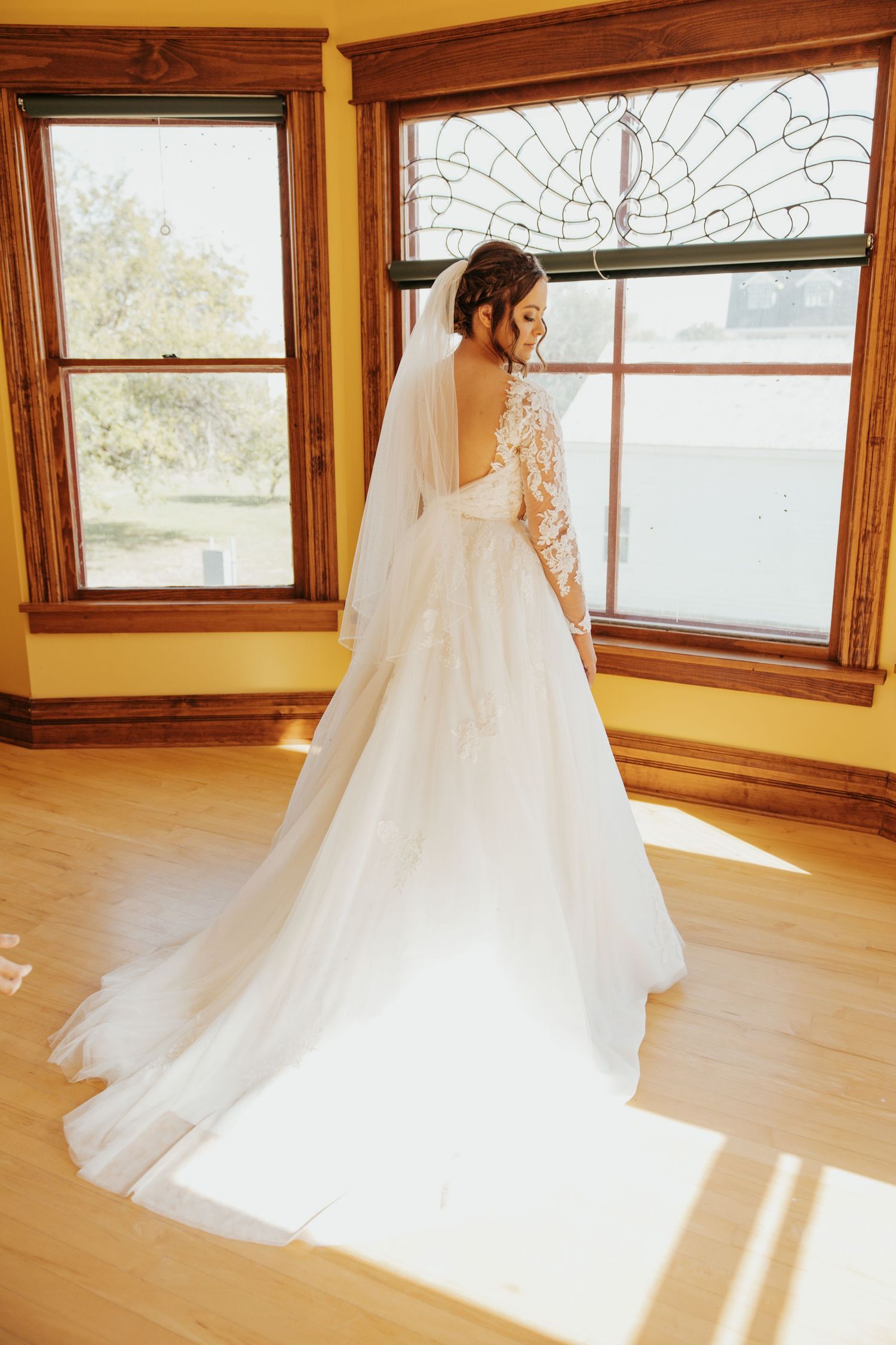 A bride in a stunning lace wedding gown stands by sunlit windows, showing off her flowing train.