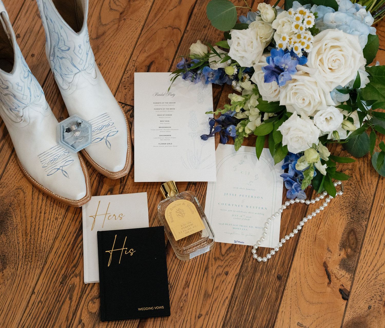 Wedding flat lay with white cowboy boots, blue and white bouquet, vow books, perfume, pearls, and invitation suite.