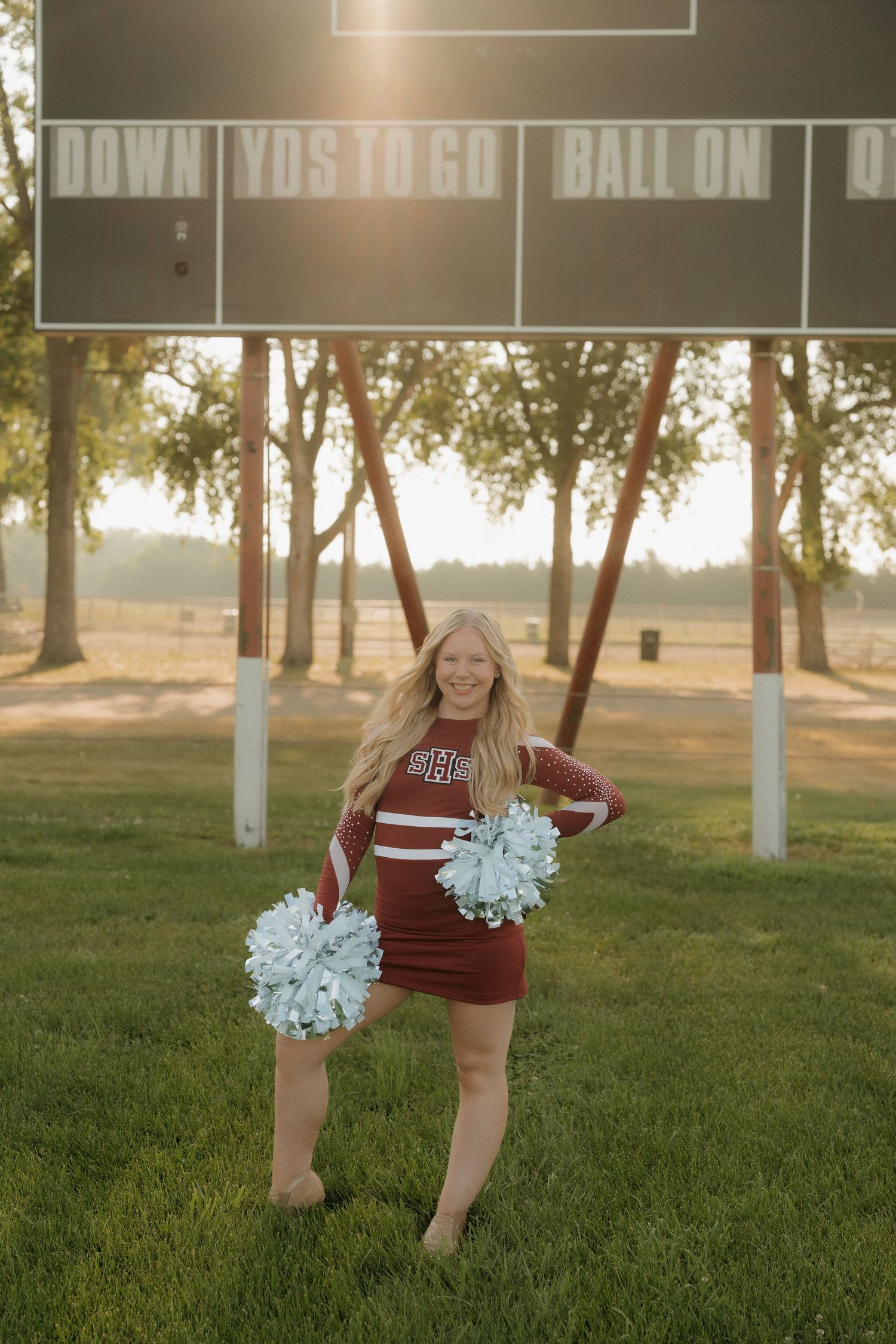 Cheerleader in red uniform with white pom-poms poses in front of a football scoreboard at golden hour.