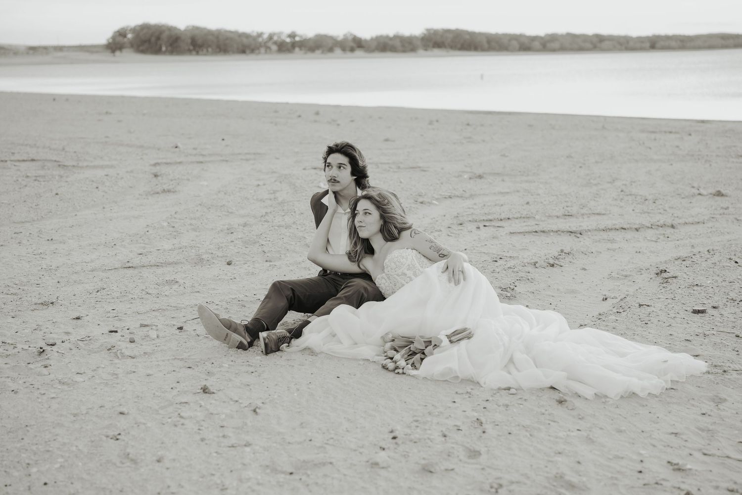 Black and white photo of bride and groom sitting together on sandy beach in wedding attire.
