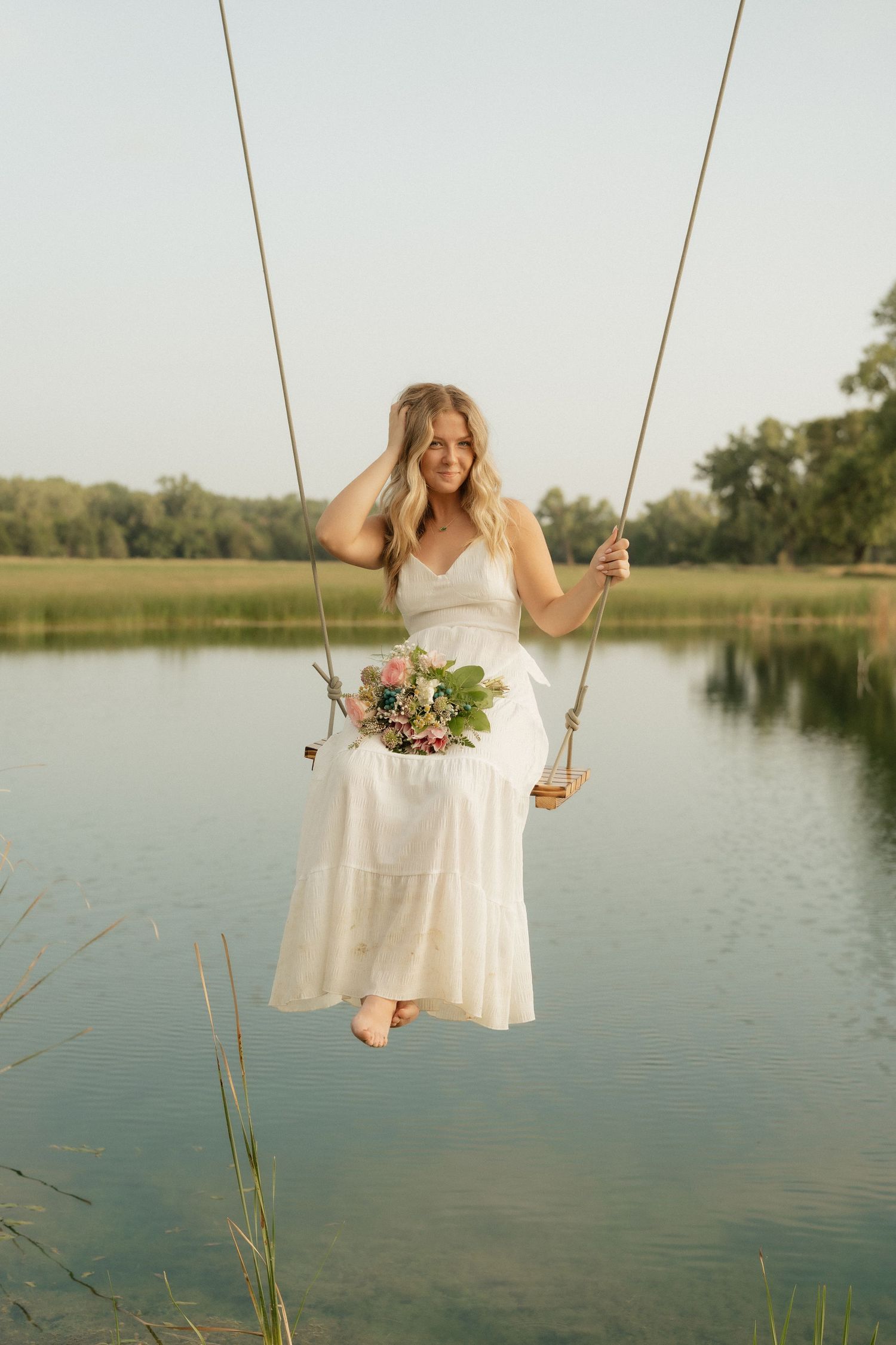 Blonde bride in white dress holding floral bouquet sits on rope swing over serene lake surrounded by green trees.