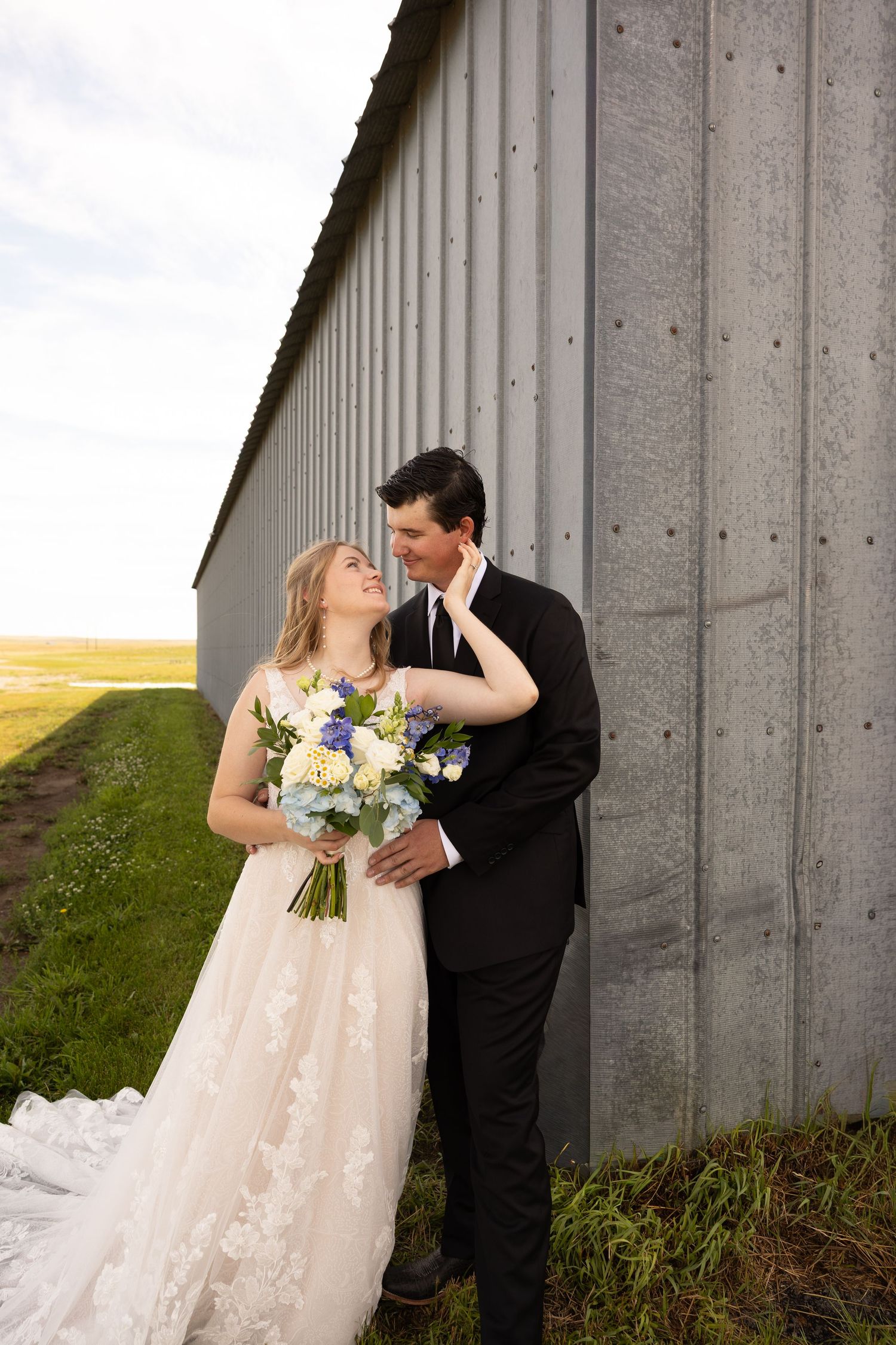 Bride and groom share a romantic moment leaning against a rustic barn wall, bride holding a blue and white bouquet.