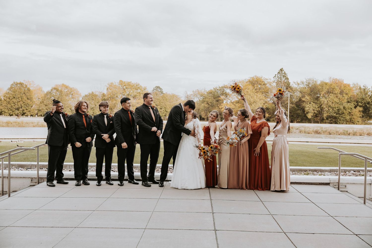 Wedding party in formal attire poses on outdoor terrace with autumn trees in background.