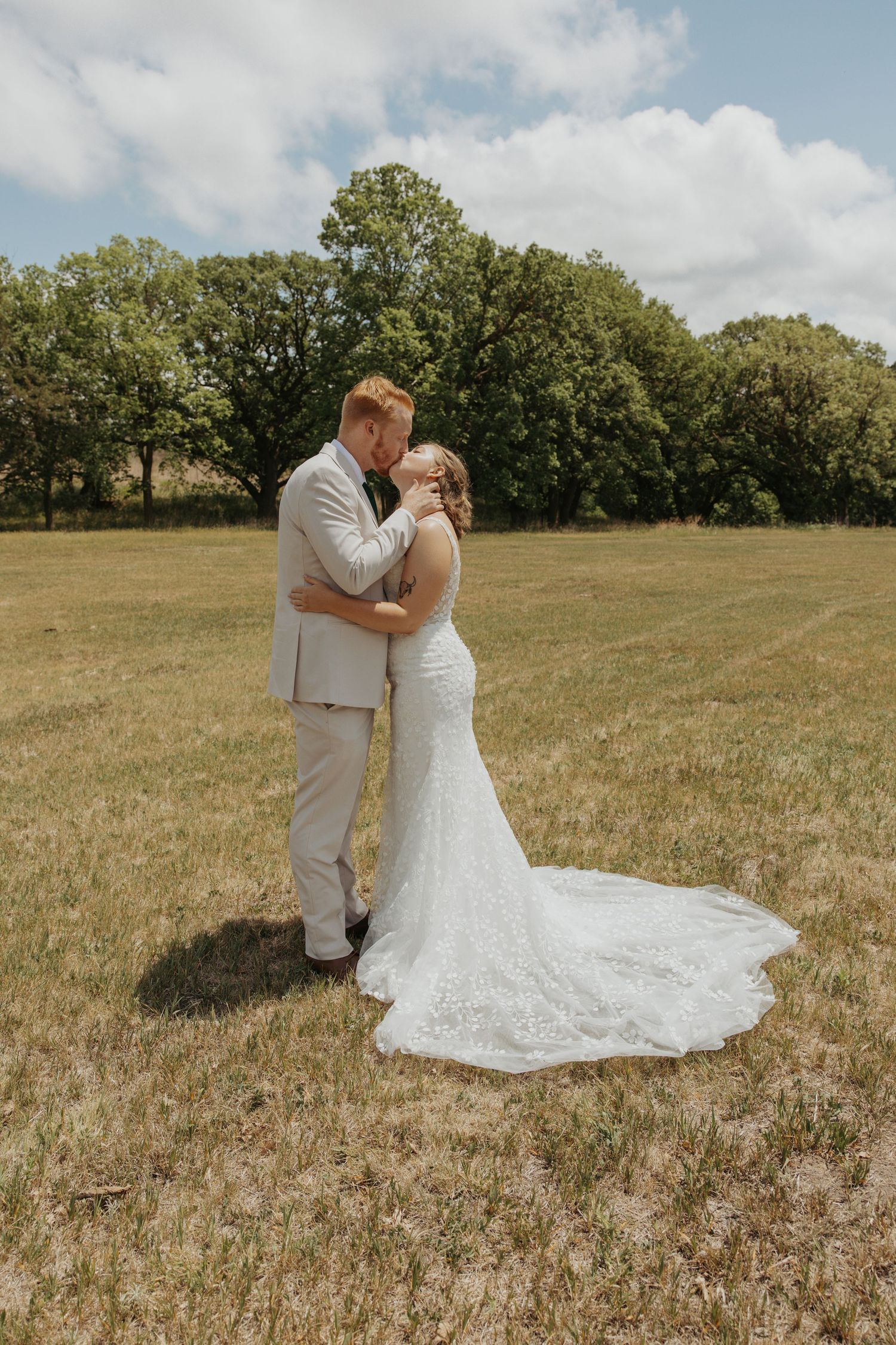 Bride and groom share a romantic kiss in a sunlit field, with lush green trees and blue skies in the background.