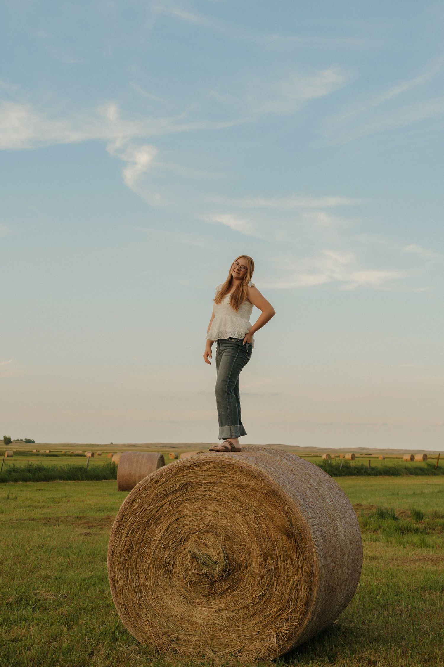 Young woman standing on a round hay bale in a green field at sunset, wearing jeans and a white top.