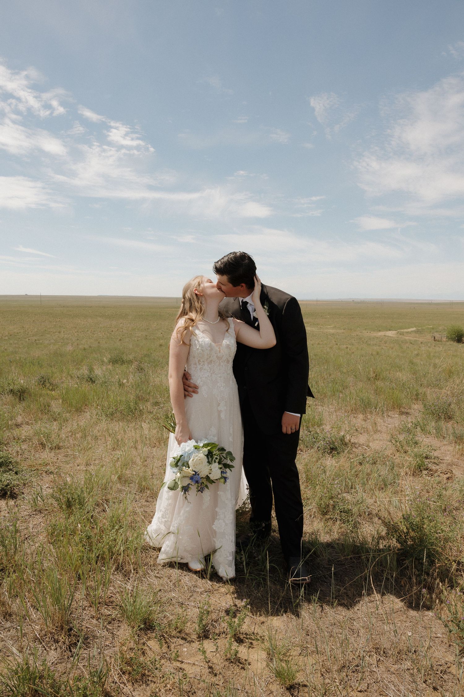 Bride and groom share a kiss in an open green field under a blue sky, bride holding a white floral bouquet.