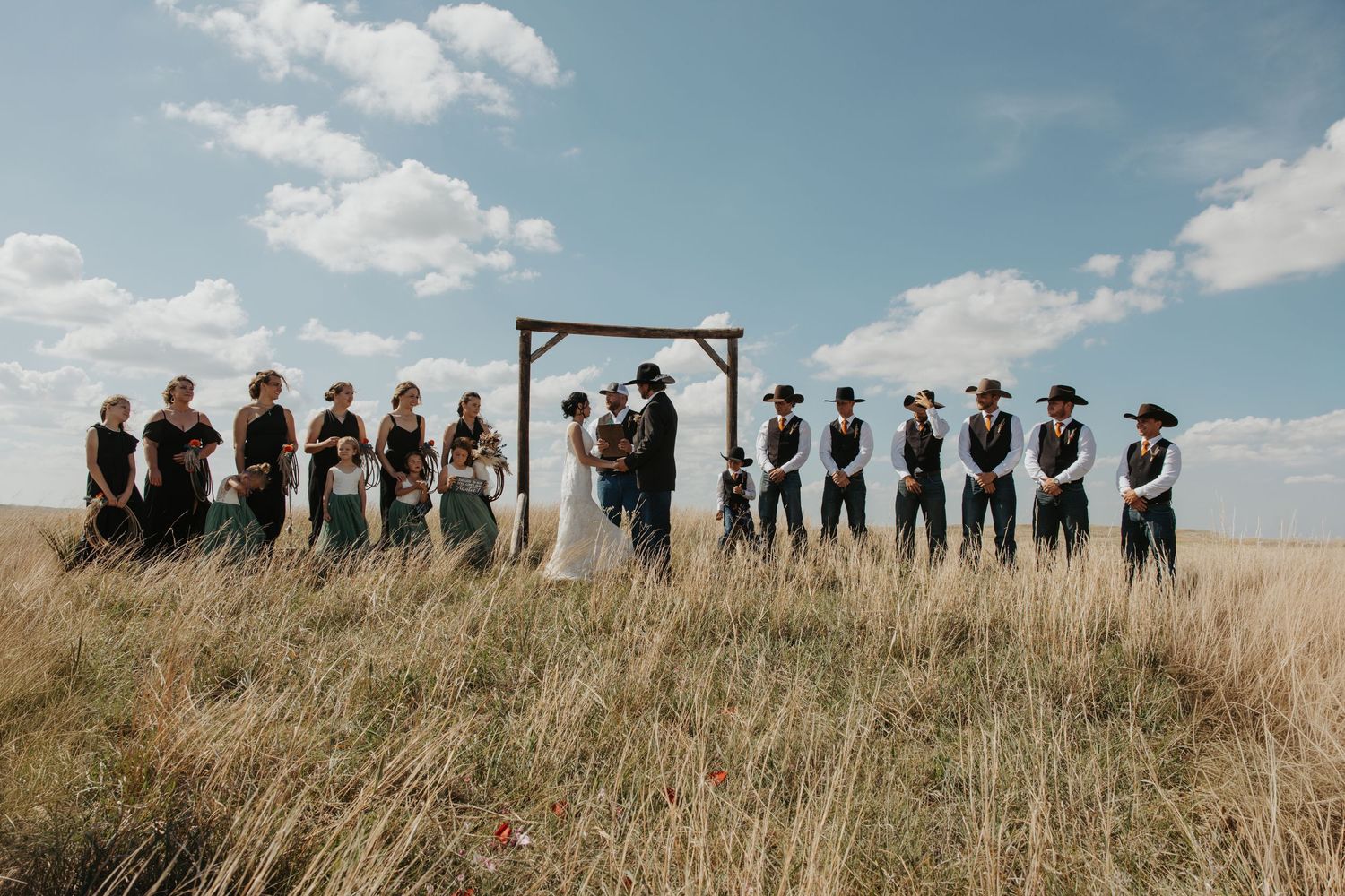 Outdoor wedding ceremony in a golden field with bridesmaids in black and groomsmen in vests under a rustic wooden arch.