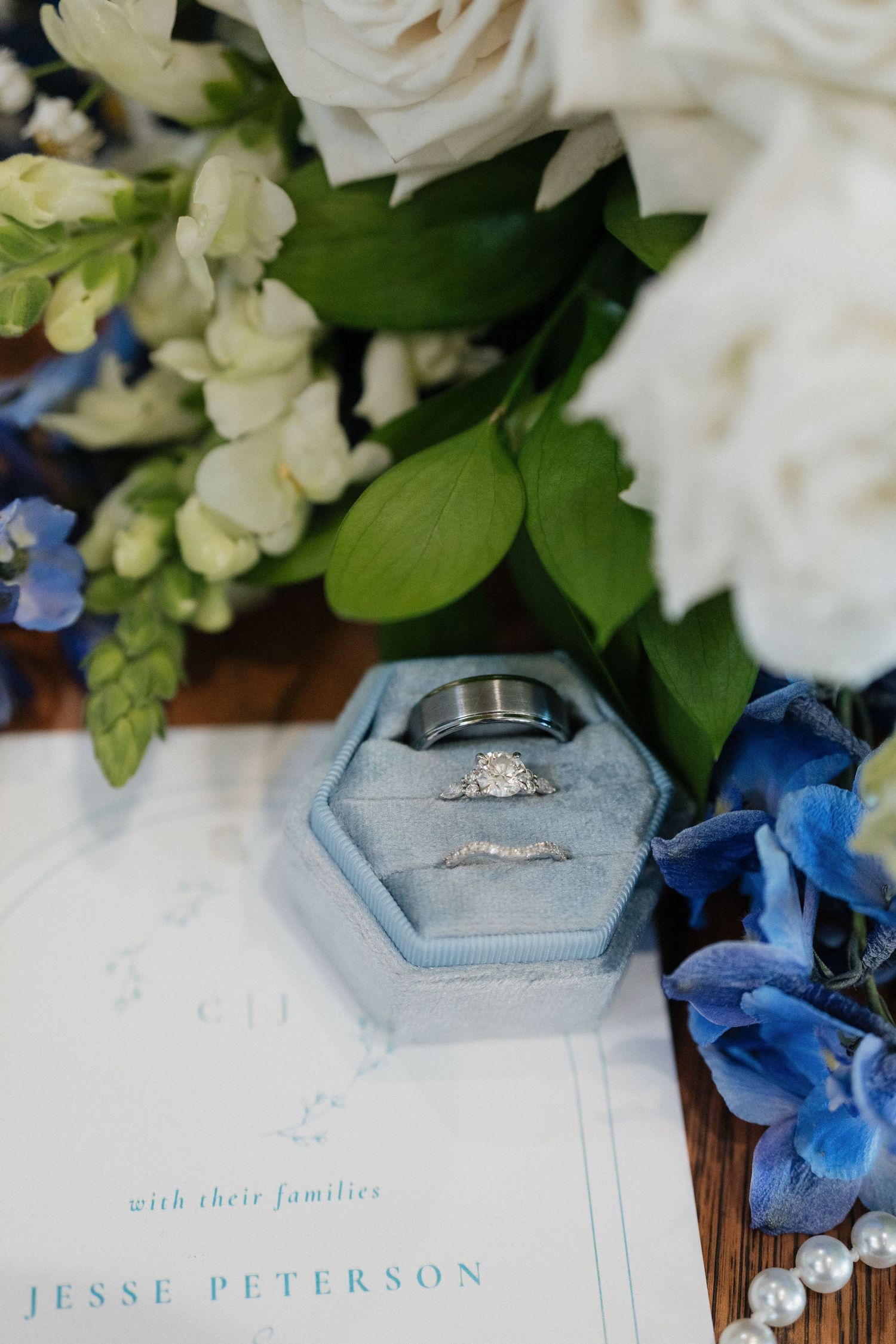 Blue velvet hexagon ring box with diamond engagement ring and wedding bands beside white and blue florals on wedding stationery.