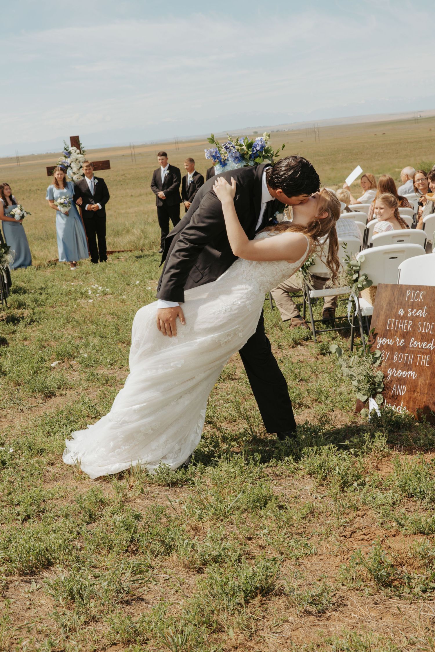 Groom dips bride for a romantic kiss at an outdoor wedding ceremony in an open field with guests watching.
