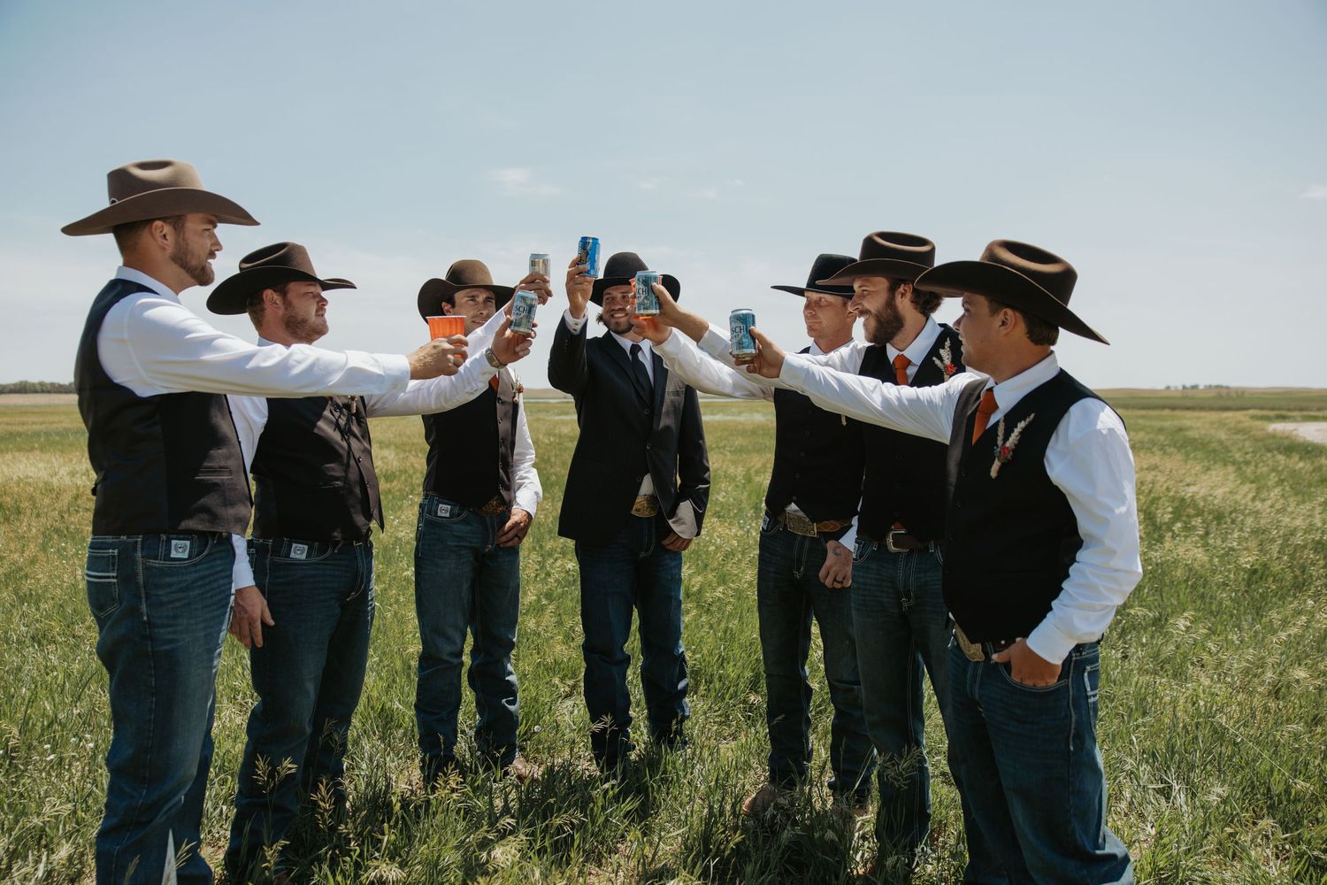 A group of groomsmen in cowboy hats and vests toast outdoors in a green field under a clear blue sky.