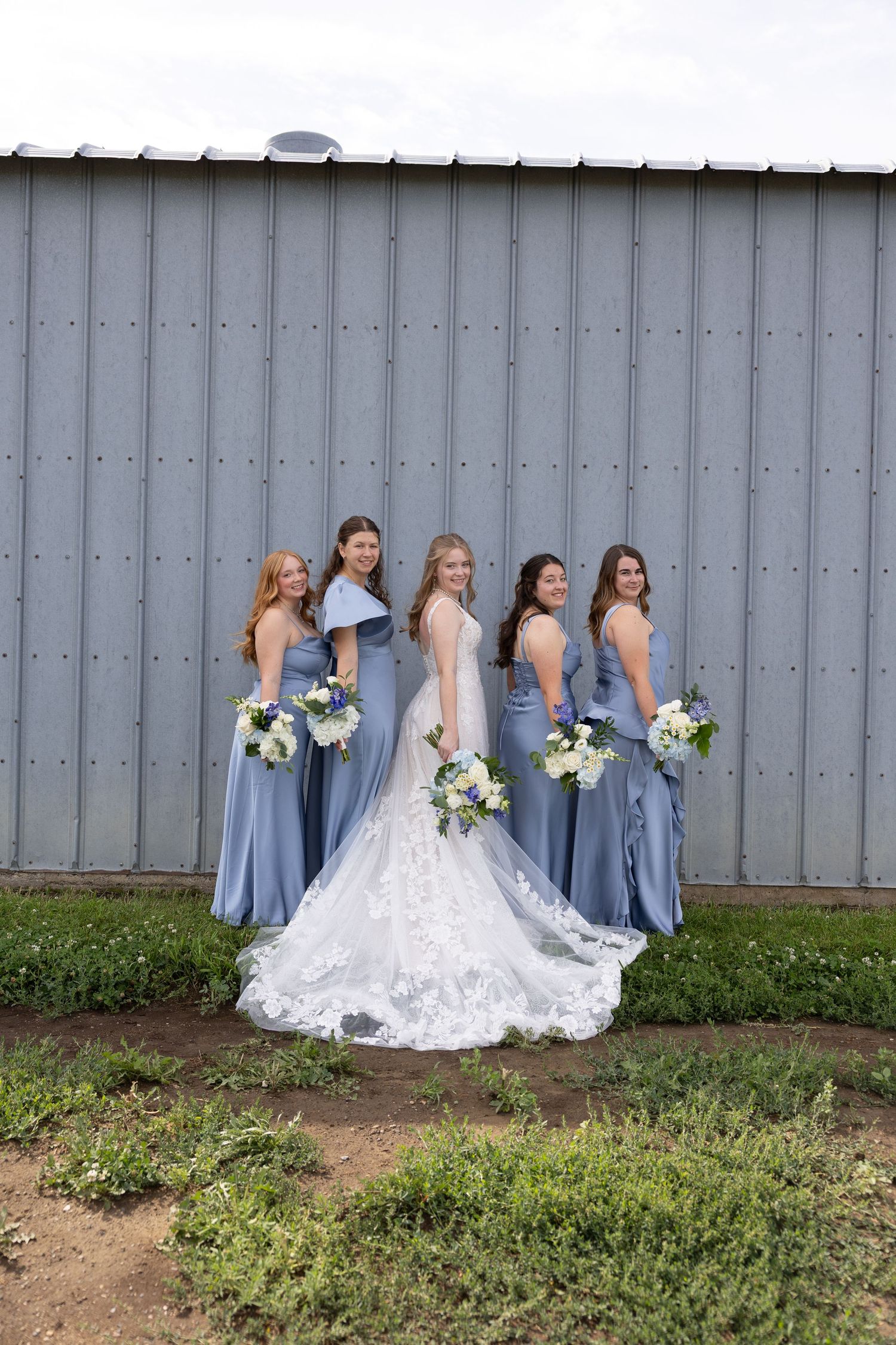 Bride in lace gown with four bridesmaids in dusty blue dresses holding white bouquets posed in front of a gray barn.