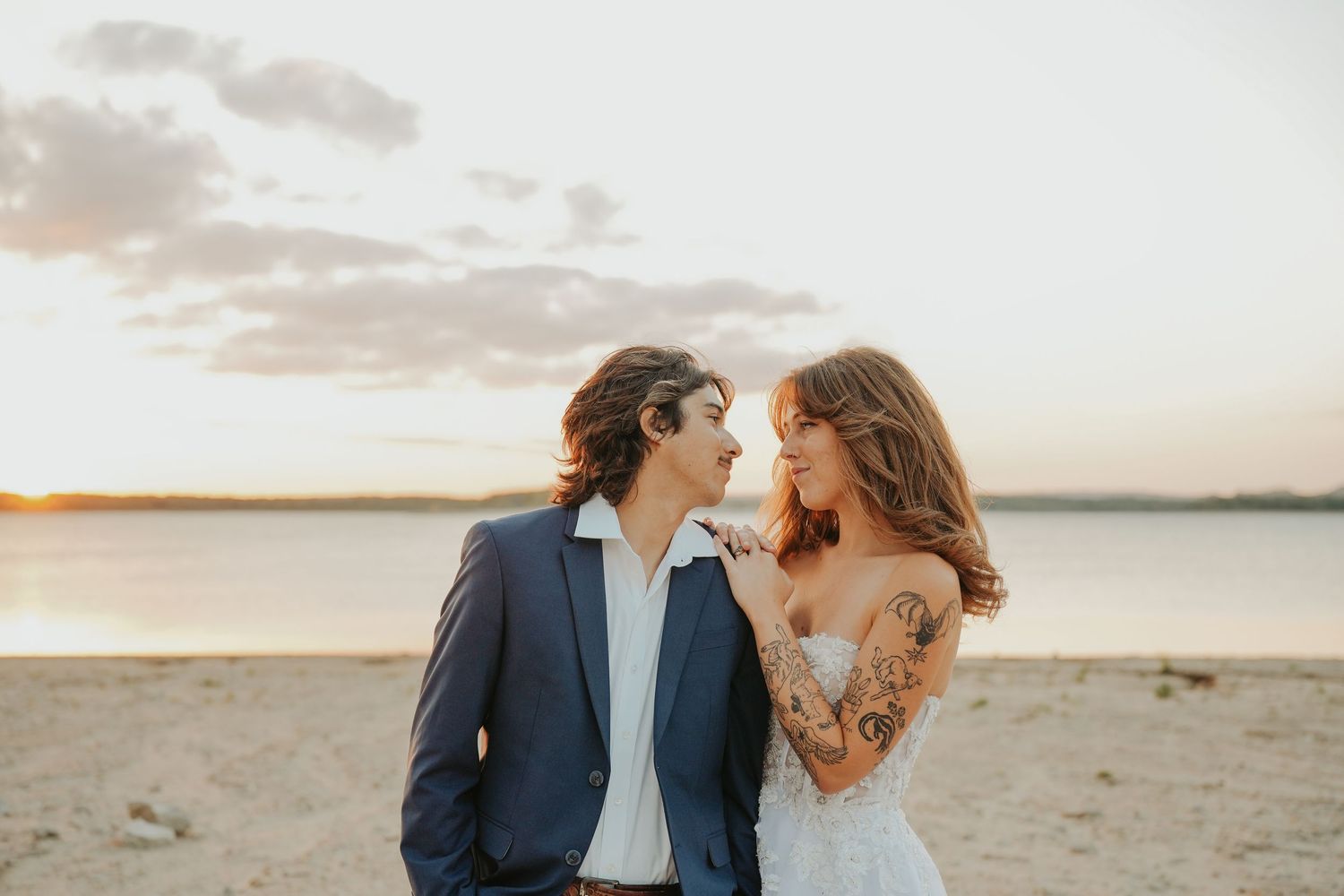 A couple in wedding attire shares a romantic moment on a beach at sunset, the bride with tattoos in a lace gown.