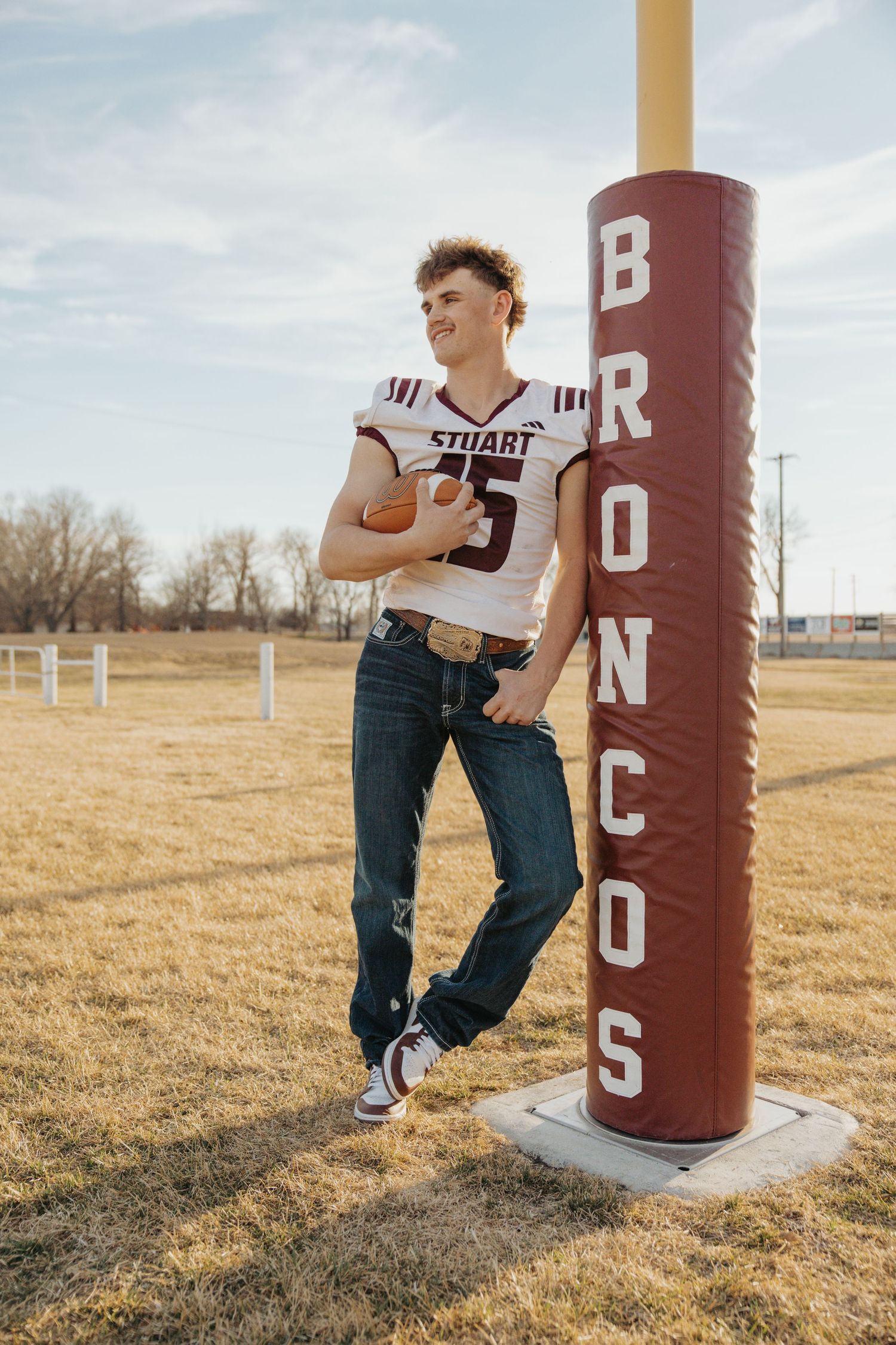 Stuart Broncos football player in uniform leaning against goalpost pad holding football on field.