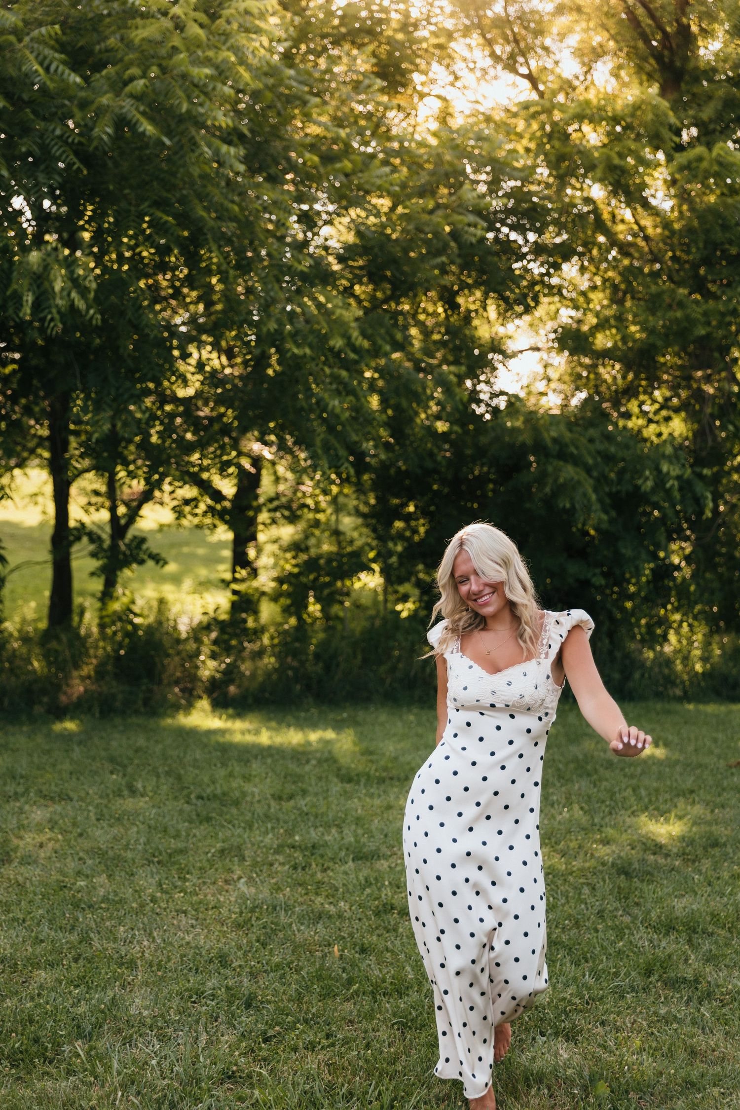 A smiling blonde woman twirls in a white polka dot midi dress in a sunlit green field surrounded by trees.