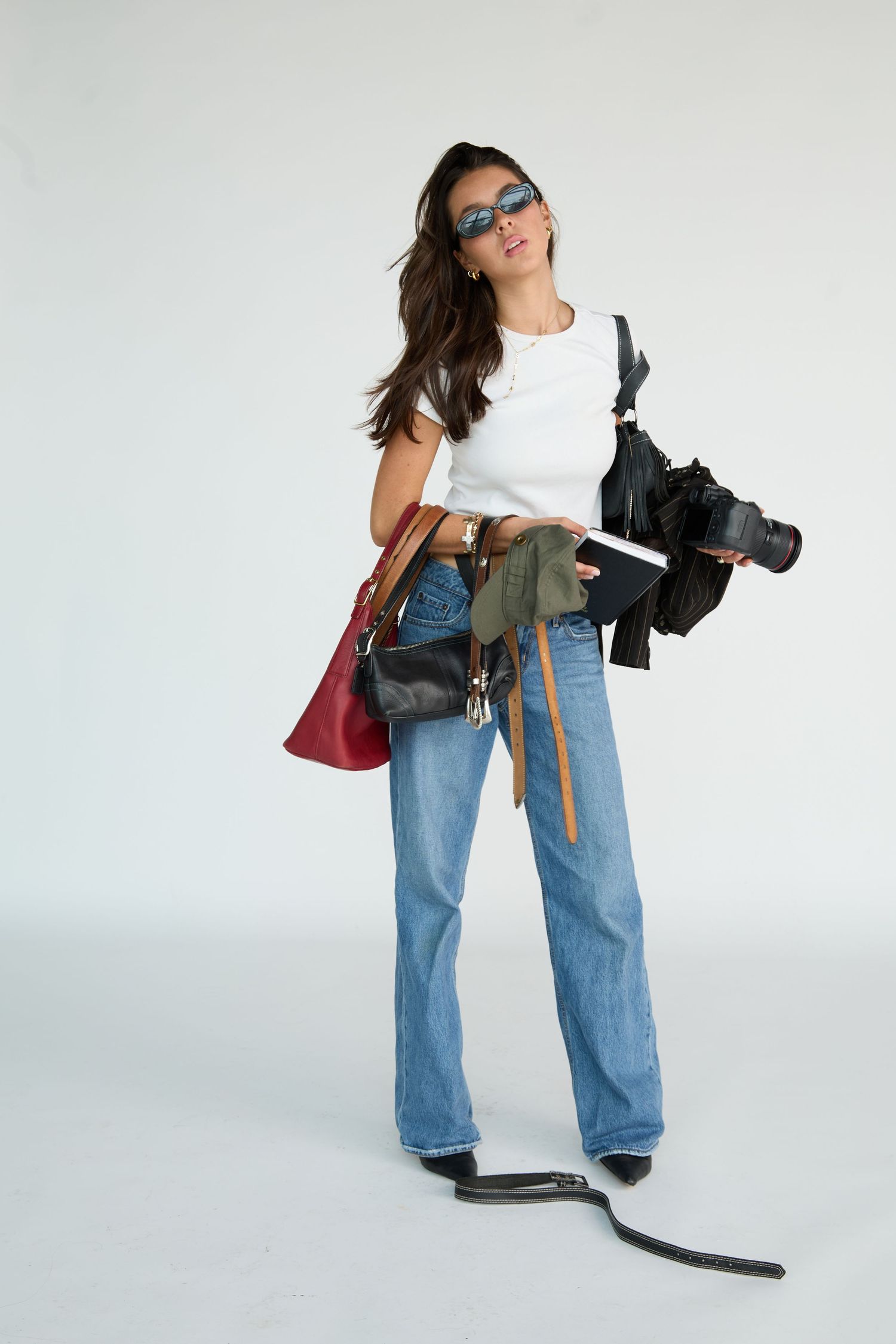 Young woman in white tee, jeans, and sunglasses holding a camera and bags in a studio setting.
