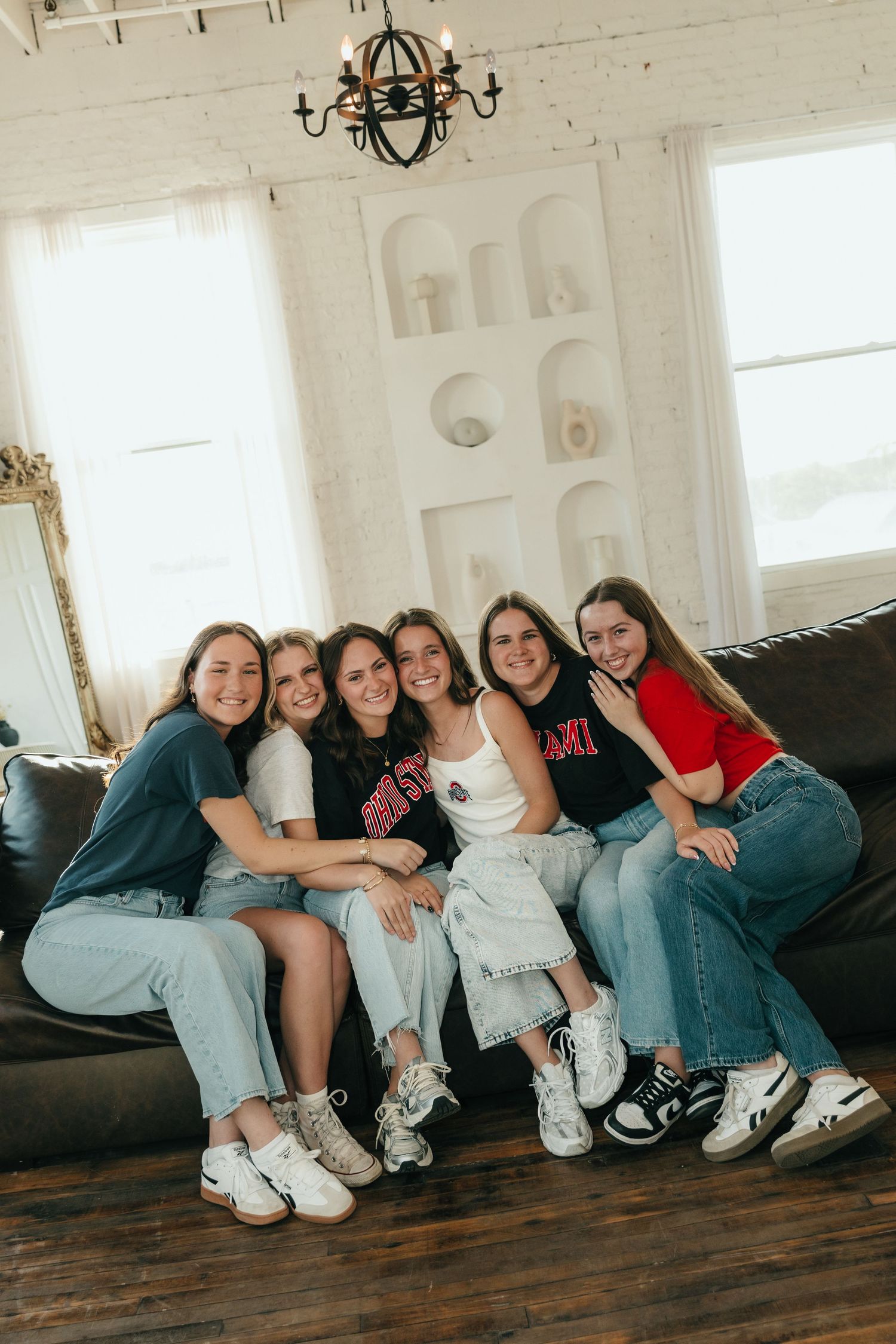 Six young women smiling and posing together on a dark sofa in a bright white living room with hardwood floors.