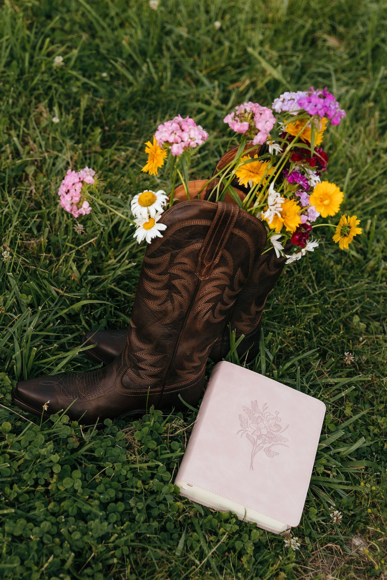 Brown cowboy boot filled with wildflowers beside a floral journal on green grass.