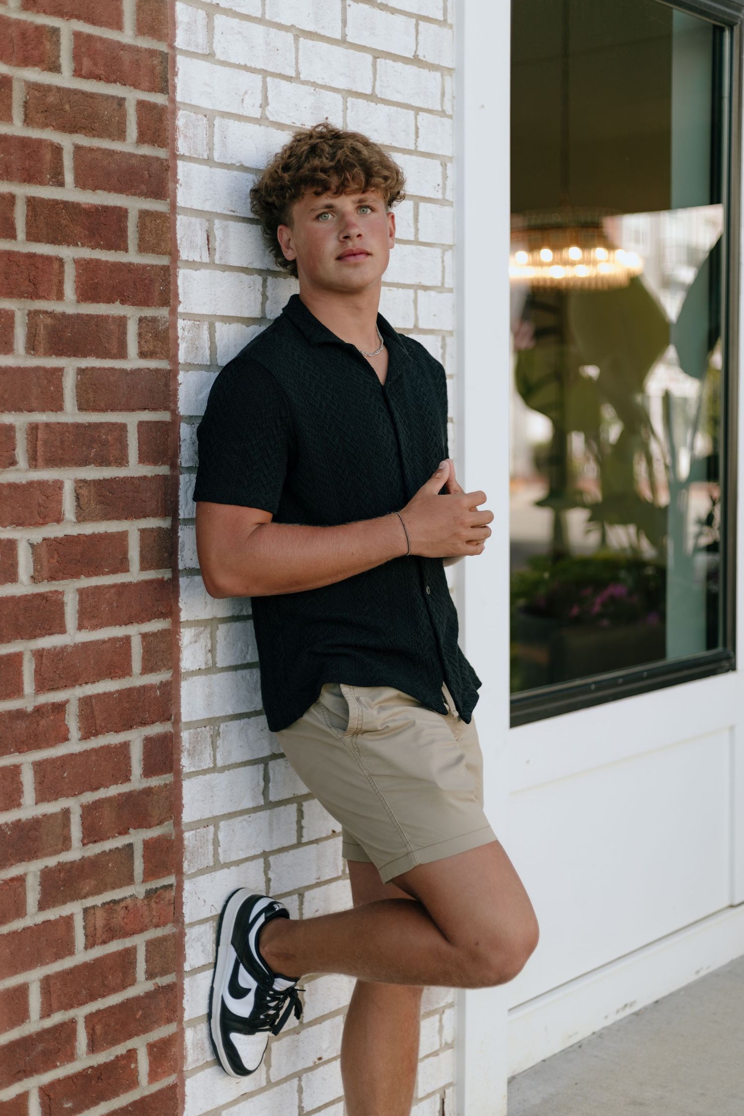Young man in black shirt and khaki shorts leaning against a brick wall outdoors.