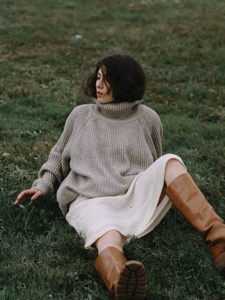 Person in cozy beige turtleneck sweater and white skirt relaxing on grass while wearing brown leather boots.