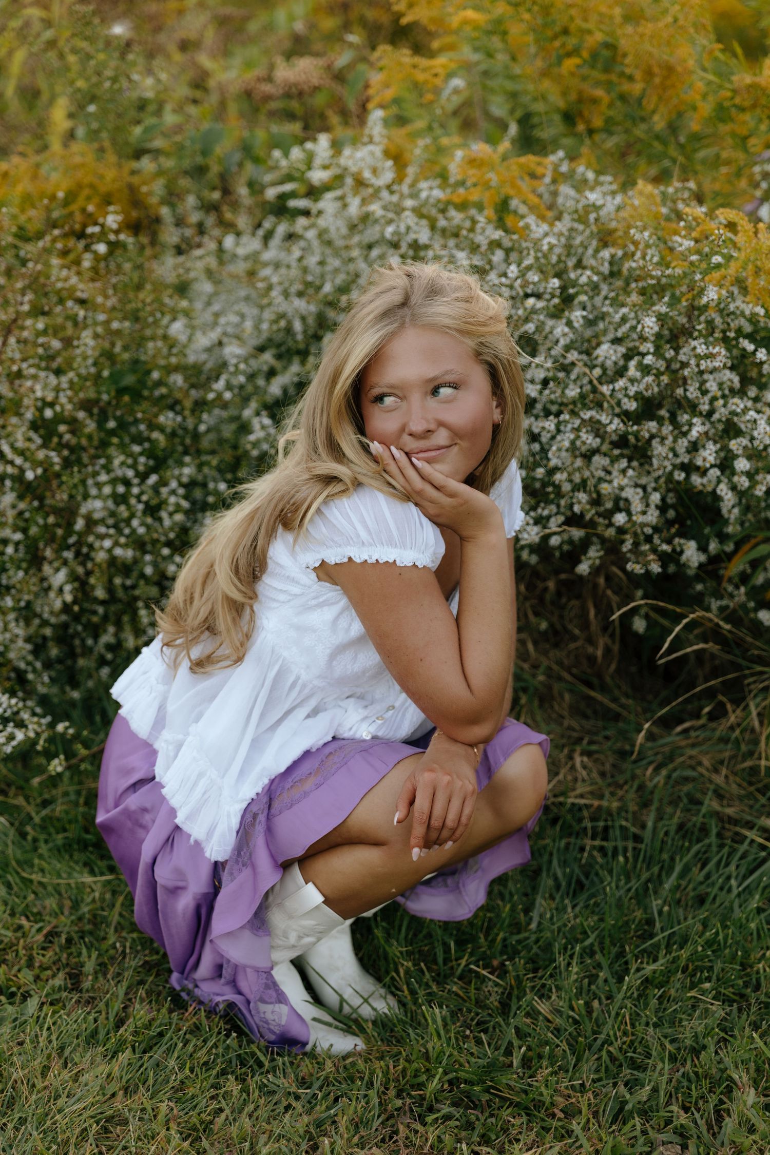 A child in a purple dress and white top sits thoughtfully in a field of wildflowers on a sunny day.