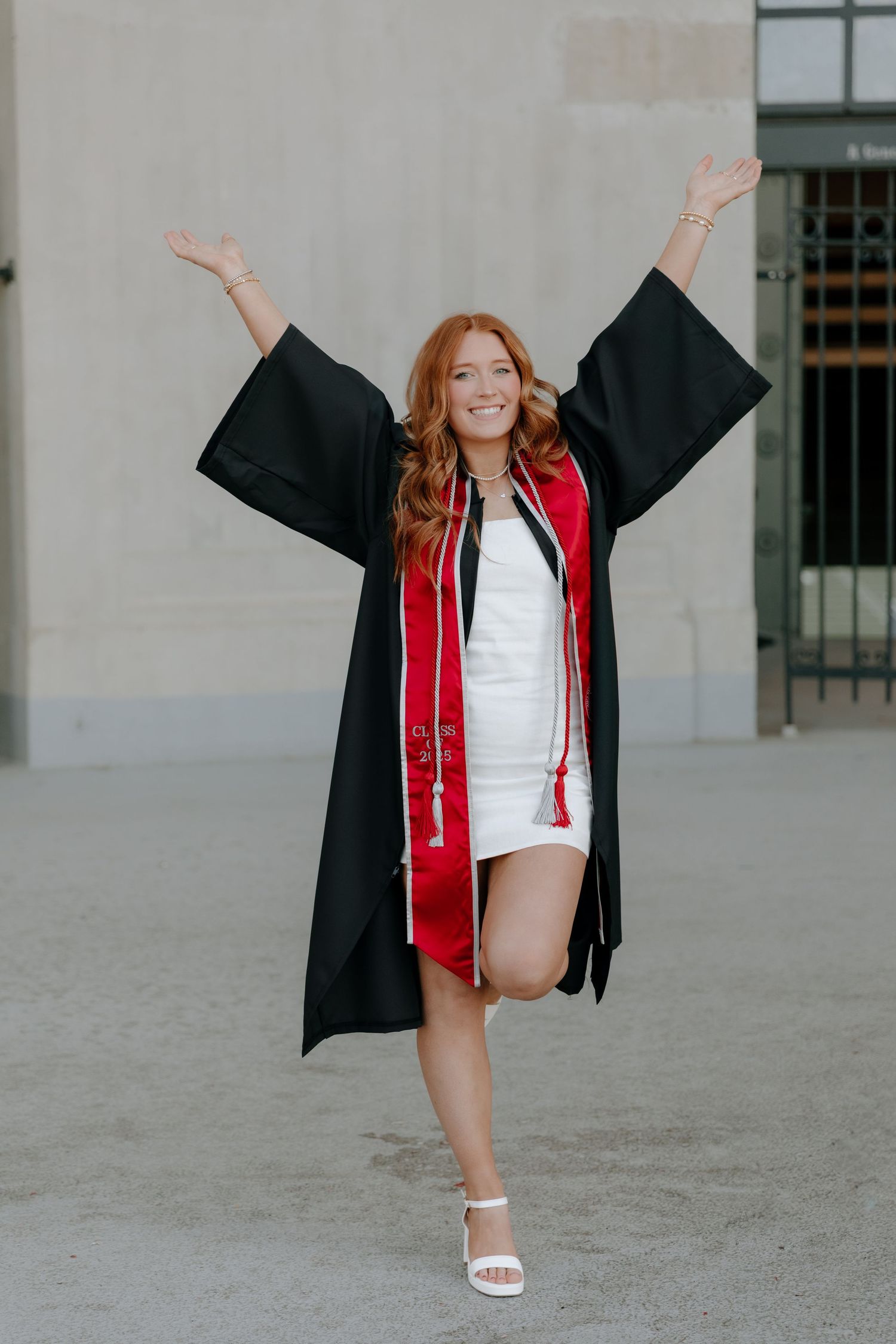Graduate in black cap and gown with red stole celebrates while wearing white dress and sandals outside building.