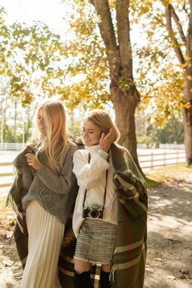 Two smiling women wrapped in a plaid blanket stand under autumn trees, one holding a vintage camera, near a white wooden fence.