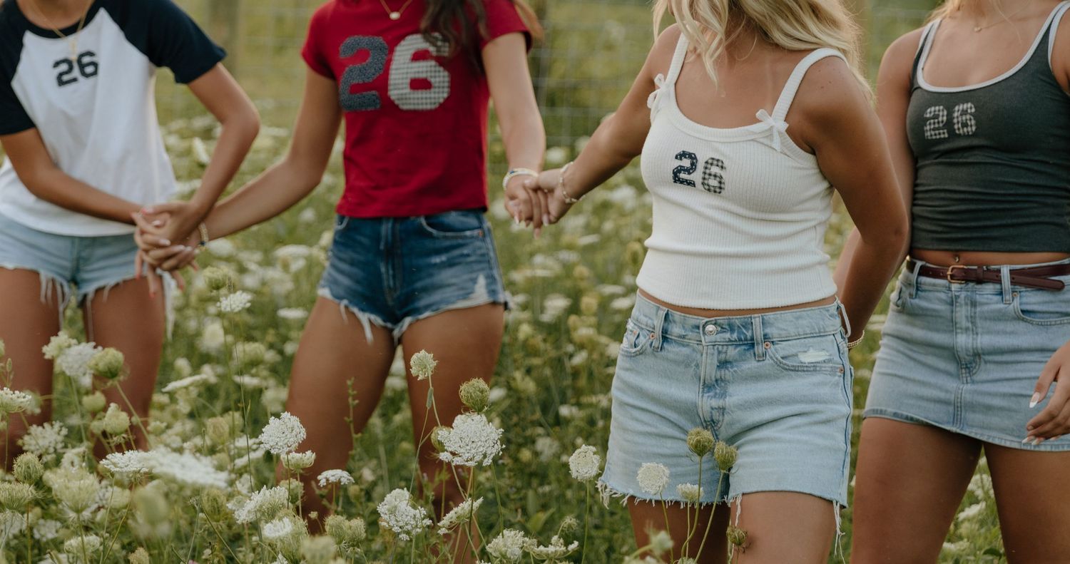 A row of people wearing summer clothing and denim shorts stand together outdoors on grass in warm sunlight.