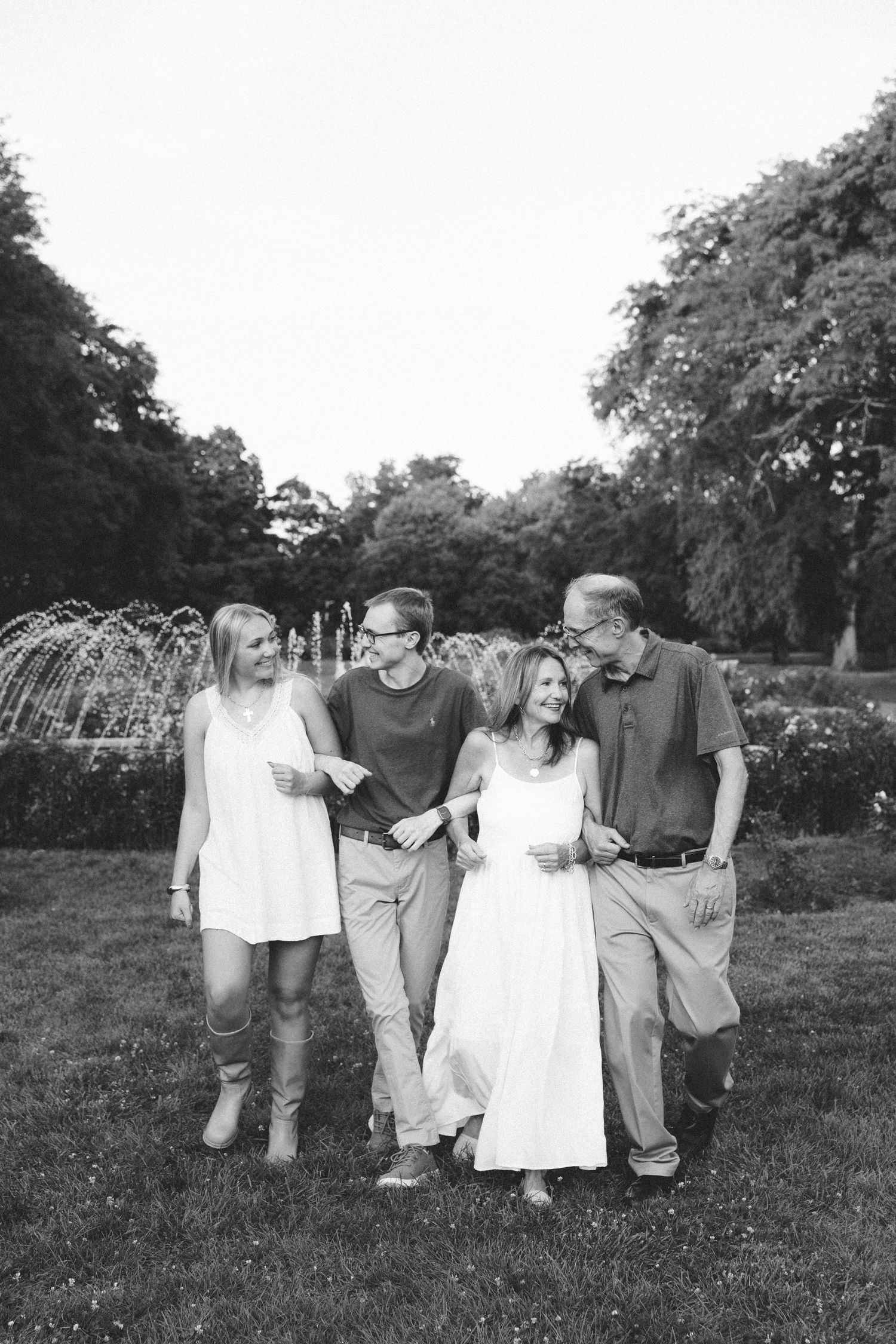 Black and white photo of two couples walking and laughing together in a lush park with a fountain backdrop.