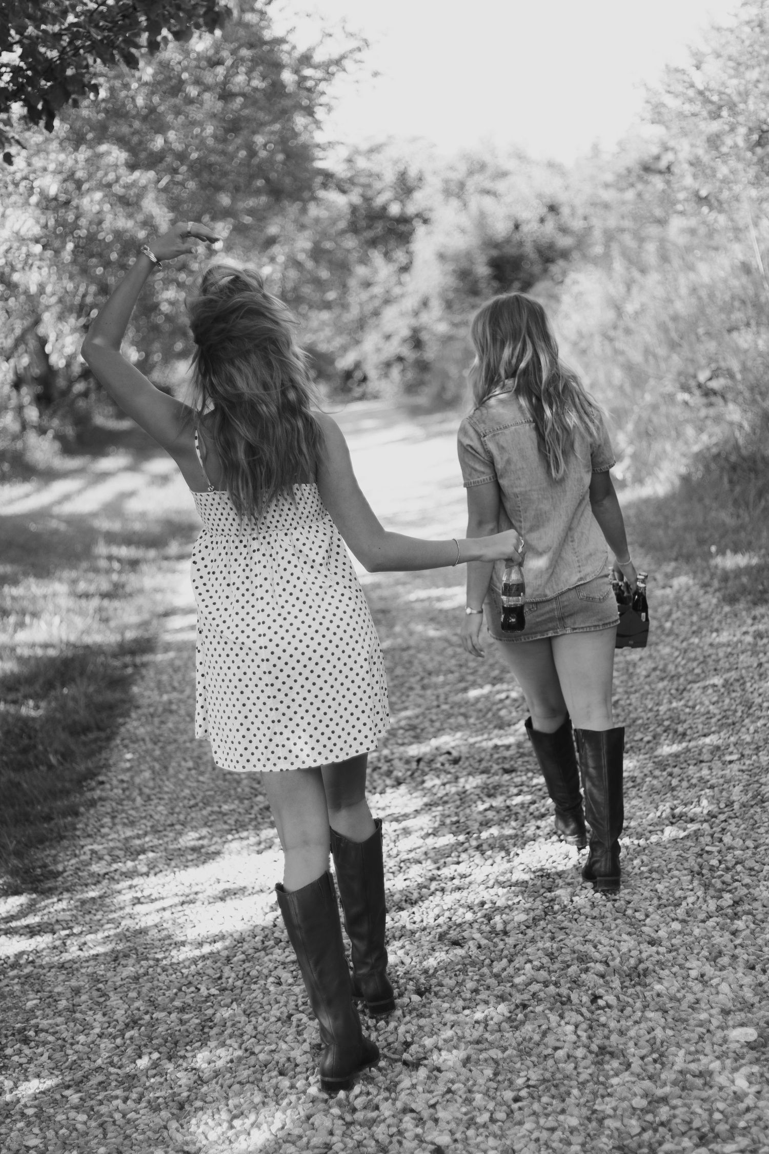 Two young women in boots and summer dresses walk down a tree-lined gravel path in this black and white photo.