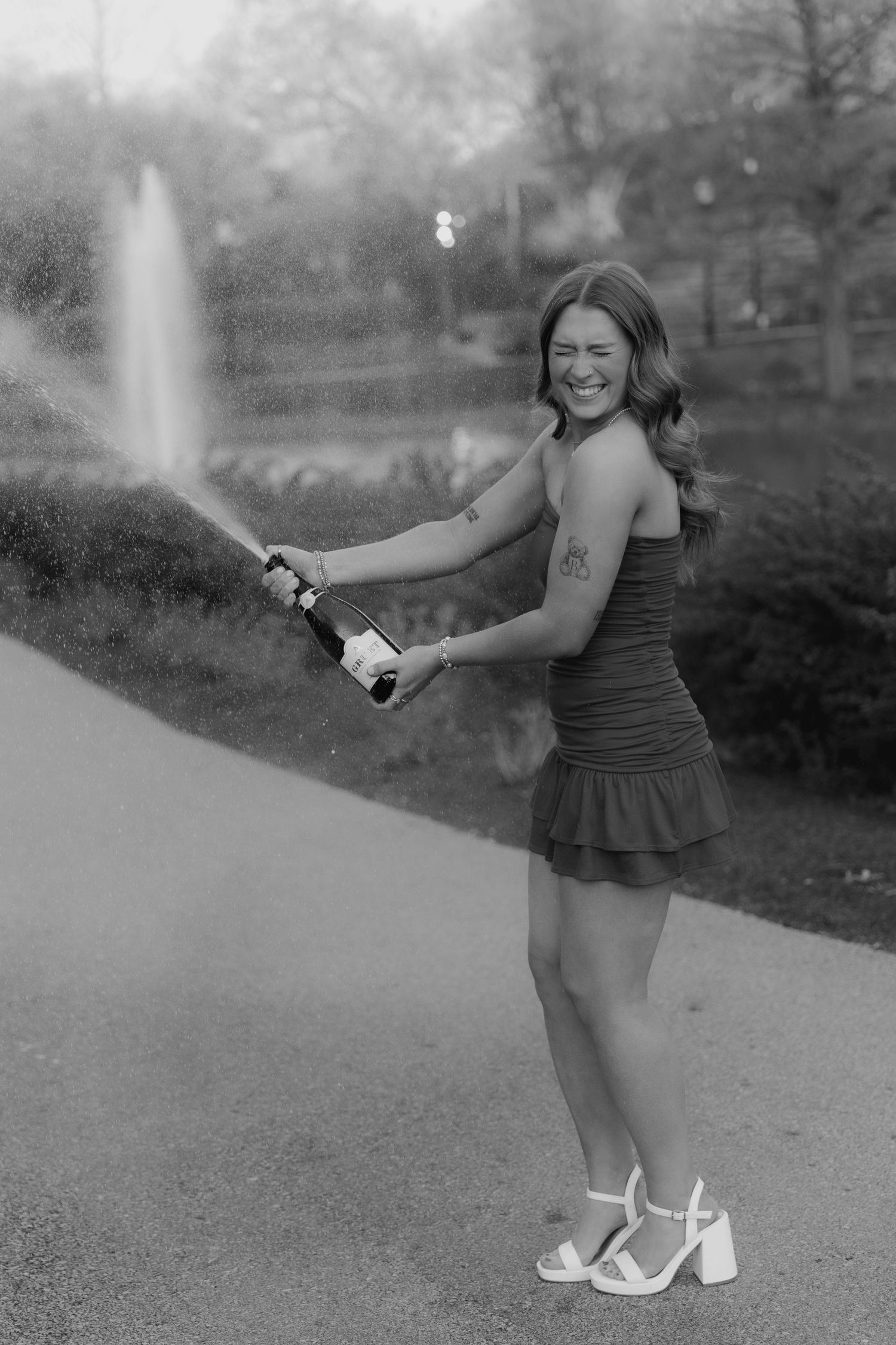 A joyful person in a black dress and platform sandals sprays champagne outdoors in black and white photography.