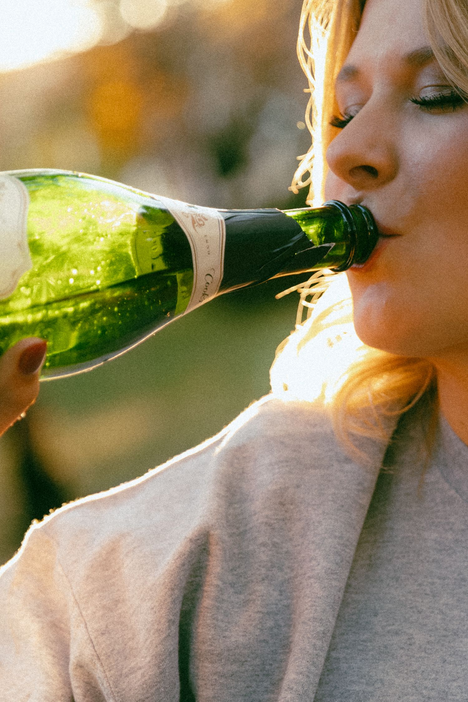 Person in grey sweatshirt drinking from green glass bottle against sunlit background.