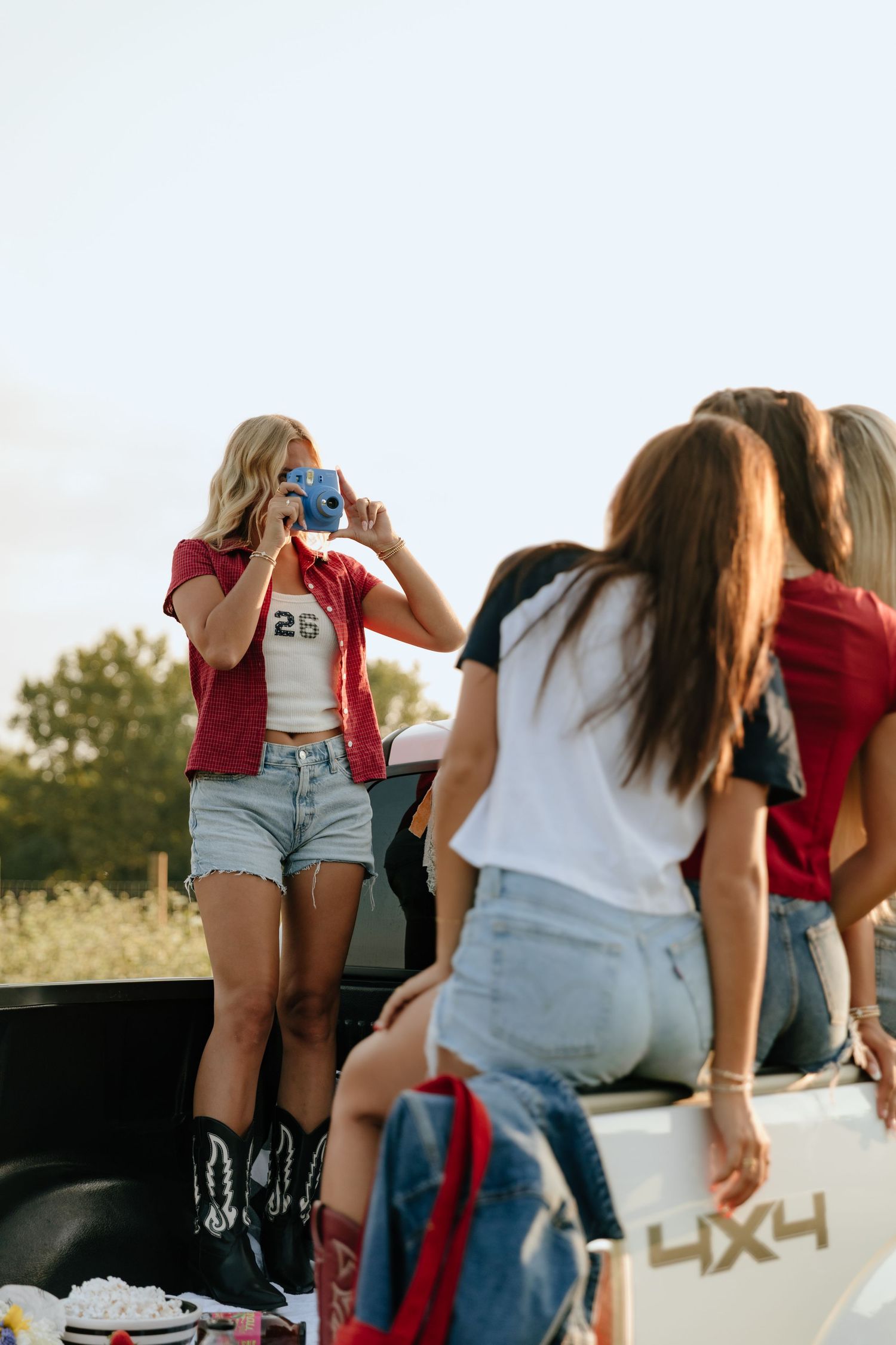 Group of friends sitting on a truck tailgate wearing casual summer outfits in golden hour lighting.