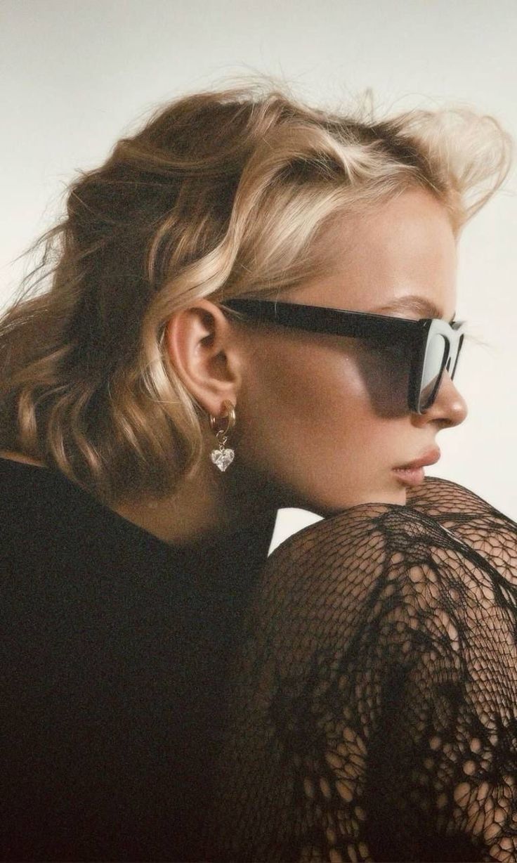 Side profile fashion portrait showing black lace top, dark sunglasses, and heart-shaped earring in moody lighting.