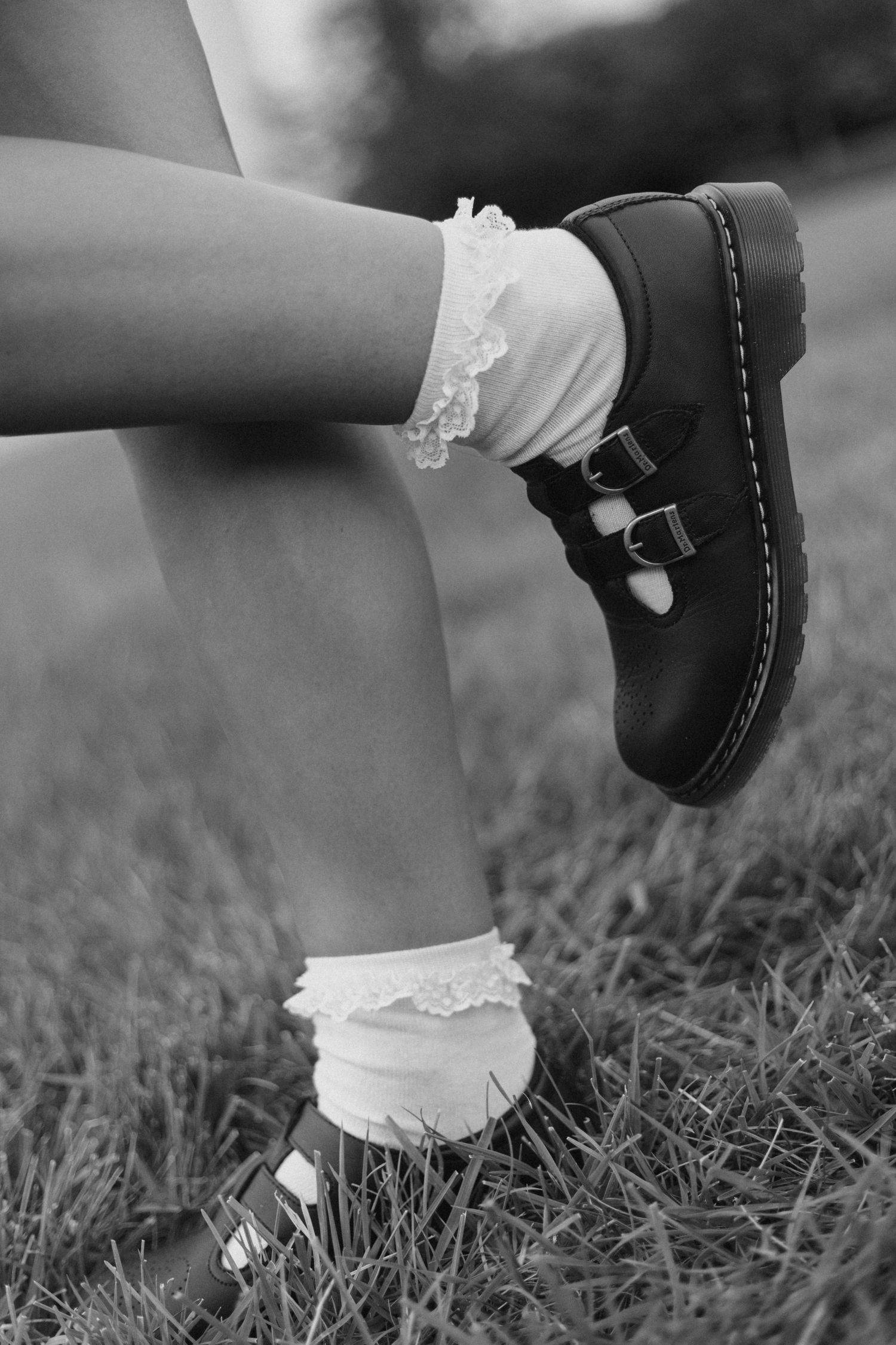 Black and white close-up of small feet wearing white socks and dark mary jane shoes on grass.