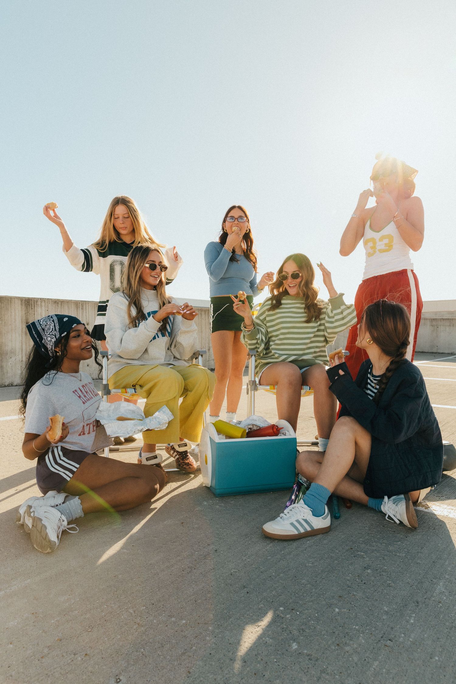 Group of young women enjoying snacks and drinks outdoors on a sunny day, sitting around a cooler.